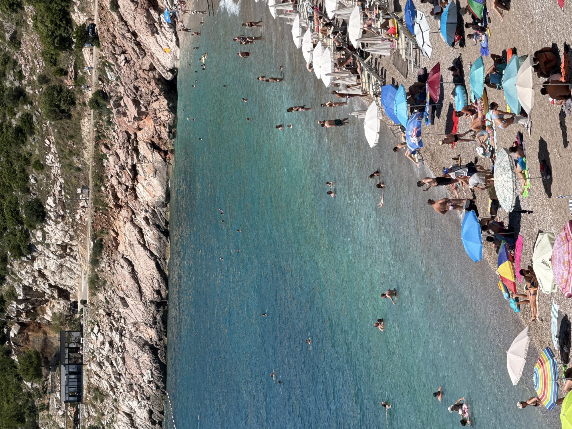 This is a high-angle, eye-level photograph of Kamenovo Beach in Kamenovo, Montenegro. The beach is characterized by its pebble shore and the clear, turquoise waters of the Adriatic Sea. Rocky, tree-covered cliffs rise steeply behind the beach, with a road and a modern, glass-fronted building visible on the hillside.
The beach is populated with numerous people engaged in various activities. Many are swimming and wading in the shallow parts of the sea, while others are sunbathing on loungers and towels on the shore. Beach umbrellas, both open and closed, are scattered across the sand, providing shade. People are seen in swimsuits and casual beach attire.
The water appears calm, with gentle ripples on its surface. The scene is bathed in bright sunlight, suggesting a warm, summer day. The overall atmosphere is one of leisure and recreation.