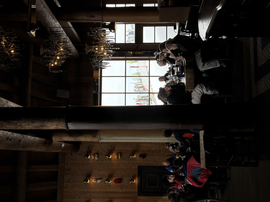 The image displays the interior of a ski lodge or restaurant in Les Allues, France. The space features dark wooden plank walls and ceiling beams, with several thick, dark wooden support columns, some wrapped in a padded material. Ornate chandeliers with multiple light sources hang from the ceiling, alongside smaller wall-mounted sconces illuminating decorative items, possibly hats or mittens, displayed on hooks along the left wall. A dark wooden door is also present on the left.
Approximately 15-20 individuals are seated at several rectangular wooden tables throughout the room. They are dressed in winter clothing, including jackets, sweaters, and some wearing beanies or ski hats. Actions include dining, drinking from glasses and bottles, and social interaction, with individuals looking at each other or at items on the table, and one person on the right viewing a smartphone.
Large, multi-paned windows dominate the background, revealing a snow-covered outdoor landscape under an overcast sky. Beyond the windows, numerous colorful skis and snowboards are vertically planted in the snow, indicative of a ski slope or equipment staging area. The floor is dark, with a patterned rug visible beneath tables on the right.