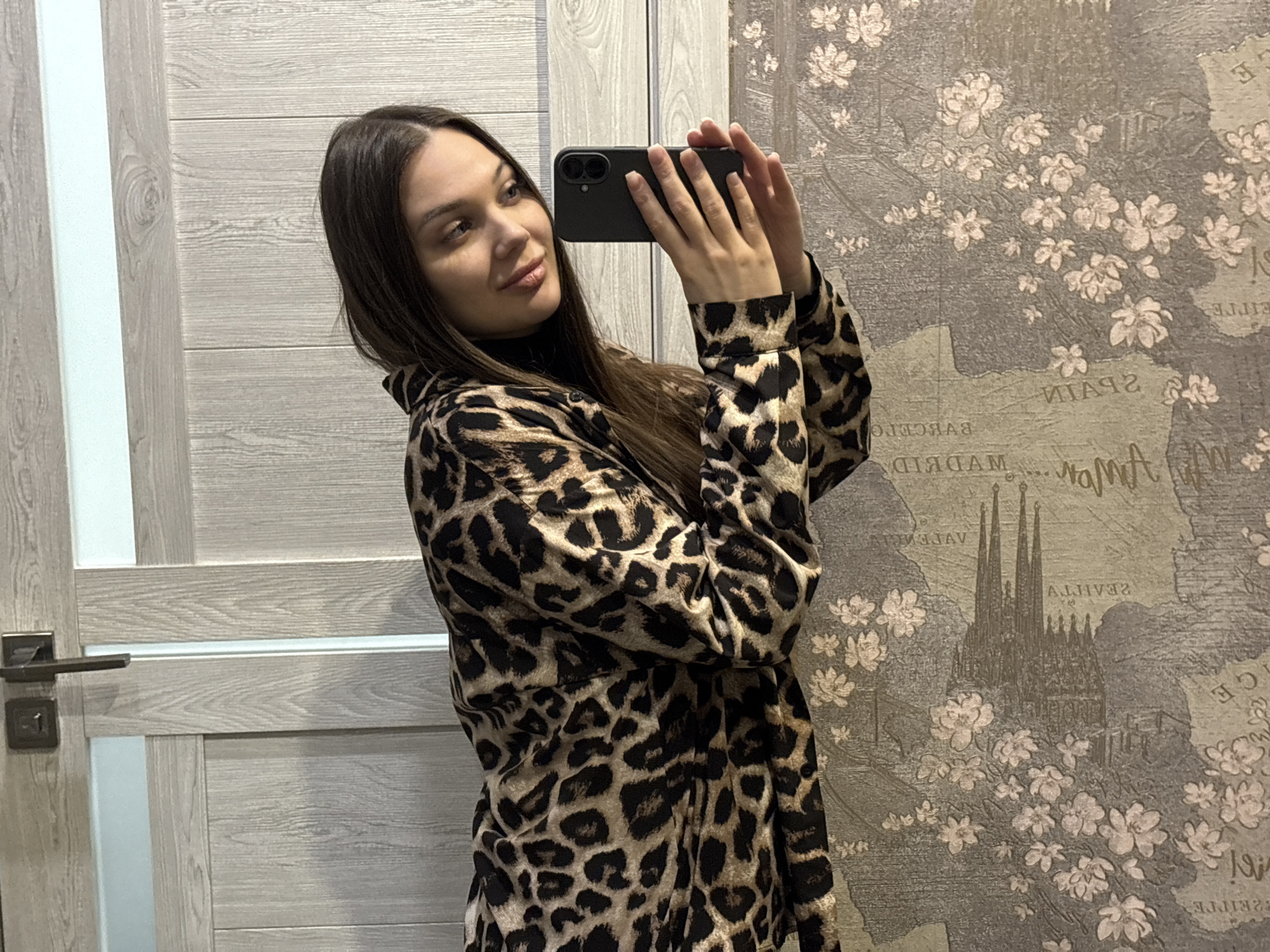 An indoor portrait captures a young woman taking a mirror selfie. She has long, straight dark brown hair, parted on the side, and a gentle, pleasant expression with a subtle smile. She wears a prominent leopard print top or jacket, possibly over a black turtleneck. She holds a dark-colored smartphone horizontally with both hands, positioned to capture her reflection, with her gaze directed slightly to her right.
The background is divided into two distinct sections. On the left, a light-colored wooden door with horizontal paneling and a vertical white accent stripe is visible. A dark metallic door handle with a keyhole is mounted on the door. On the right, the wall is covered with decorative wallpaper featuring a vintage-style map pattern overlaid with numerous light-colored floral designs. Due to the mirror reflection, text on the map appears reversed. Clearly visible reversed place names include "SPAIN," "MADRID," "VALENCIA," and "BARCELONA." A detailed illustration of the Sagrada Familia in Barcelona is also prominently featured on the wallpaper. The scene is brightly lit, indicative of indoor lighting. The activity takes place in what appears to be a residential or private interior, located in Yekaterinburg, Russia.