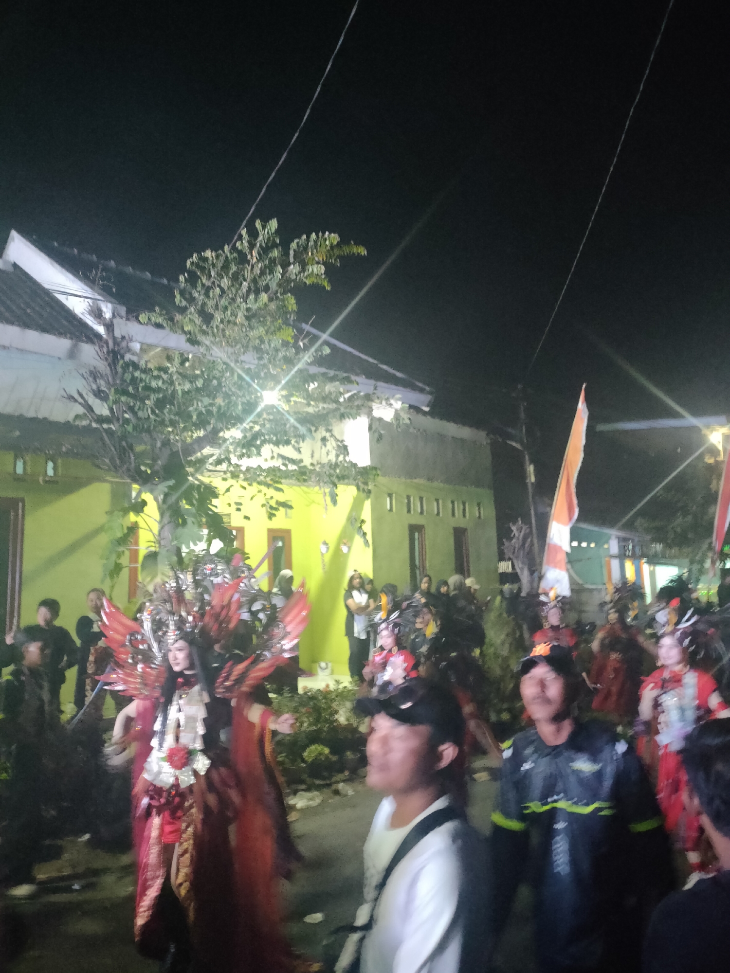 The image depicts a nighttime scene in Tongaswetan, Indonesia, likely during a cultural event or parade. In the foreground, several people are visible. A woman in an elaborate, red and silver costume with large feathered wings and an intricate headdress is the central figure. She has long, dark hair and is looking to her left. To her right, a man wearing a black baseball cap and a dark jacket with neon green accents is looking upwards. Further to the right, another person in a white shirt is partially visible. In the background, a group of people in similar elaborate costumes, some with feathered headpieces, are also present. An Indonesian flag, red and white, is prominently displayed, attached to a pole. The setting appears to be a street lined with houses, one of which is painted bright yellow. Trees with sparse foliage are scattered throughout the scene, and overhead power lines are visible against the dark sky, illuminated by bright lights. The overall atmosphere suggests a lively celebration.