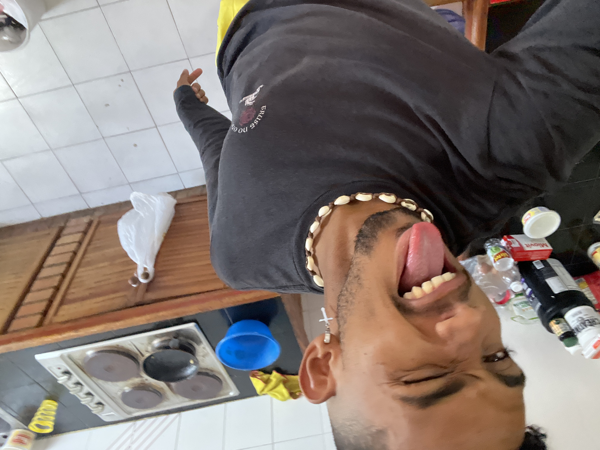 The scene is an indoor kitchen located in Porlamar, Venezuela. A male individual, positioned centrally, faces the camera at a slightly elevated angle. He has dark hair styled with a fade, a mustache, and a short beard. His right eye is closed in a wink, and his tongue is extended. He wears a dark long-sleeved t-shirt with a graphic on the left chest that includes a rose, hands, and the text "TRUST NO ONE." A necklace made of cowrie shells is visible around his neck, and a silver hoop earring with a cross pendant is present on his right earlobe. A segment of yellow apparel is discernible at the bottom of the frame. The background features a kitchen counter with a black surface, holding a silver electric stovetop equipped with four circular burners. A small black pot rests on one of the stovetop burners. Adjacent to the stovetop, a blue plastic bowl and a crumpled yellow fabric cloth are visible on the counter. To the left, a white countertop displays various items, including a white container labeled "CREATINE," a dark bottle, a red box marked "Miovit," and a clear plastic bottle. The wall above the counter is covered with white tiles. Below the counter, a wooden base cabinet with a textured surface features a hook from which a white plastic bag hangs. The floor is composed of white square tiles, exhibiting scattered dark debris.