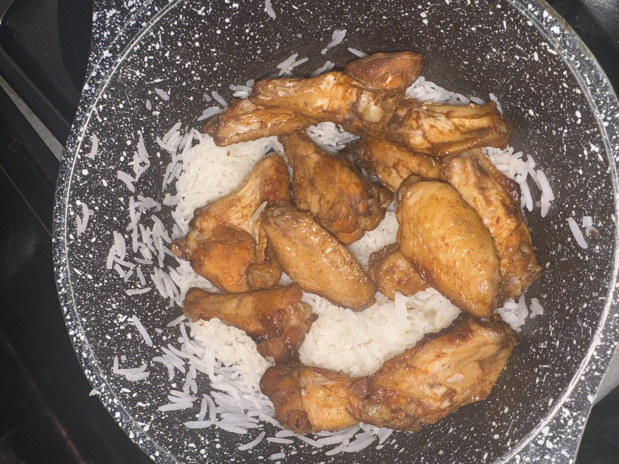A close-up, overhead shot shows chicken wings nestled in a bed of white rice in a speckled black pan. The chicken wings are golden brown and appear to be coated in a glossy sauce. The rice is fluffy and fills the center of the pan, with some grains spilling over the edges. The pan is positioned on a dark, indistinct surface, possibly a stovetop. The lighting is dim, casting a slight shadow. The scene is static, with no discernible activity. The image was taken in an indoor setting, likely a kitchen.