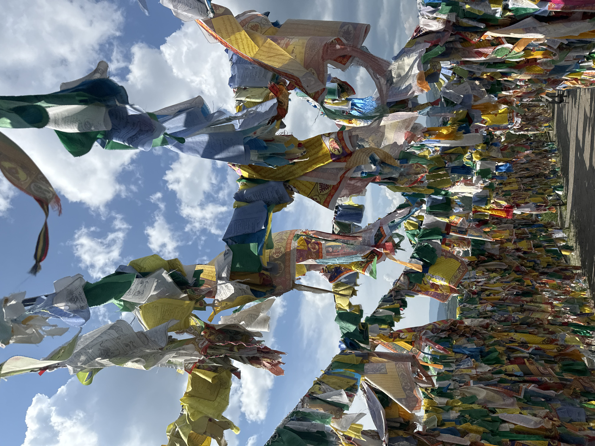 A dense collection of colorful prayer flags, predominantly white, blue, yellow, green, and red, are strung across poles against a bright blue sky with fluffy white clouds. The flags are tattered and weathered, suggesting they have been exposed to the elements for some time. Many of the flags bear printed symbols and text. The prayer flags are arranged in multiple layers, creating a vibrant canopy. Below the flags, a path or road is visible, flanked by more prayer flags attached to a fence or structure, forming a tunnel-like effect. In the distance, rolling hills can be seen under the cloudy sky. This scene is characteristic of the Ivolginsky Datsan, a Buddhist monastery located near Ulan-Ude, Russia, where such prayer flags are a common sight. There are no people visible in the image.