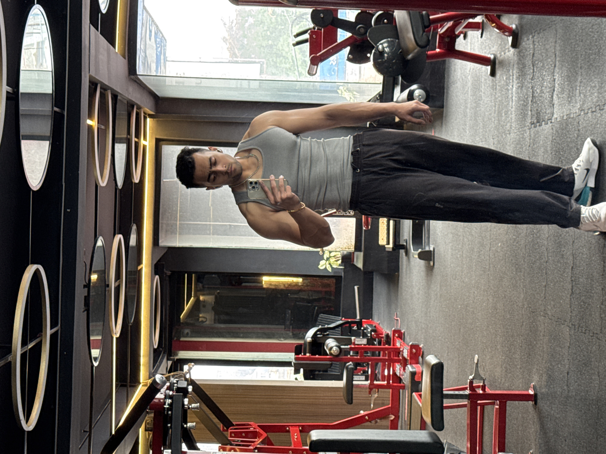 A man is captured in a mirror selfie inside a modern gym in Kolkata, India. He stands upright, facing slightly away from the mirror, holding a smartphone with his left hand to take the picture. He wears a fitted grey tank top, dark black trousers or track pants with the word "One" faintly visible, and white Nike sneakers with a black swoosh and light blue accent. A silver chain is visible around his neck, and he wears a gold bracelet on his left wrist. His eyes appear to be partially closed or looking downwards, focusing on his phone or posing for the shot.
The gym features a dark aesthetic with bright red gym equipment. In the foreground, parts of a red and black seated weight machine are visible. Other red gym machines are scattered throughout the background. The floor is covered with dark grey interlocking rubber mats. Along a dark wall, a series of oval-shaped mirrors with light-colored frames are arranged horizontally, possibly with integrated backlighting, creating bright accents. Large windows along one side of the gym allow natural light to filter in, suggesting it is daytime. The overall setting is well-lit and appears to be a contemporary fitness center.