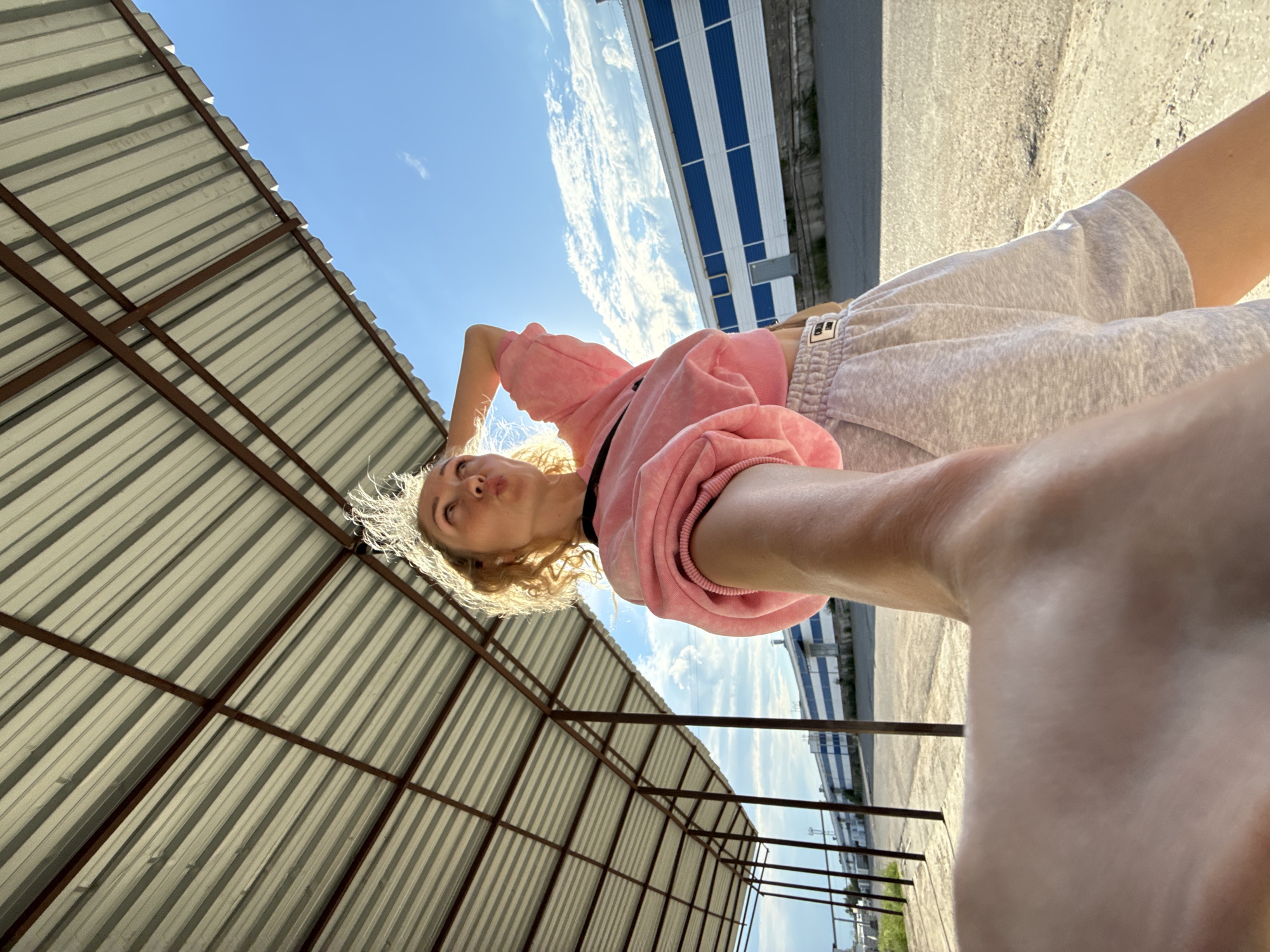 A young woman with blonde curly hair is posing for a selfie outdoors under a metal roofed structure. She is wearing a pink cropped sweatshirt and grey shorts. Her left arm is raised, and her hand is resting on her head. The background shows a blue sky with white clouds, a long building with blue and white stripes, and a dirt ground. The location is identified as Chaikovsky, Russia. The lighting suggests it is daytime, likely sunny.