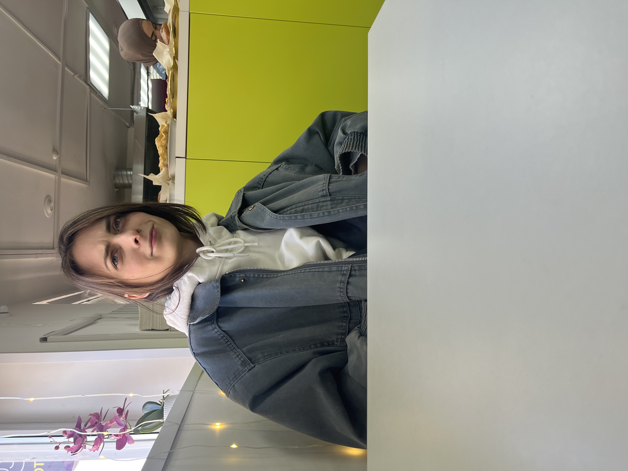 An indoor, vertically oriented image features a young woman seated at a white table, looking directly at the camera. She has shoulder-length brown hair and is wearing a white hooded sweatshirt under a dark grey or charcoal-colored jacket. Her expression is neutral with a slight gaze of curiosity.
The setting appears to be a casual eatery, cafe, or cafeteria. A prominent bright lime-green wall is visible behind her. Above a counter along this wall, another person, wearing a light brown head covering, is partially visible, seemingly attending to various pastries or baked goods displayed on the counter.
The ceiling features typical commercial fluorescent strip lighting. In the bottom left corner of the frame, a potted orchid plant with purple blooms is adorned with warm white fairy lights, adding a decorative touch. The large white table in the foreground takes up a significant portion of the right side of the image. No visible text is present.