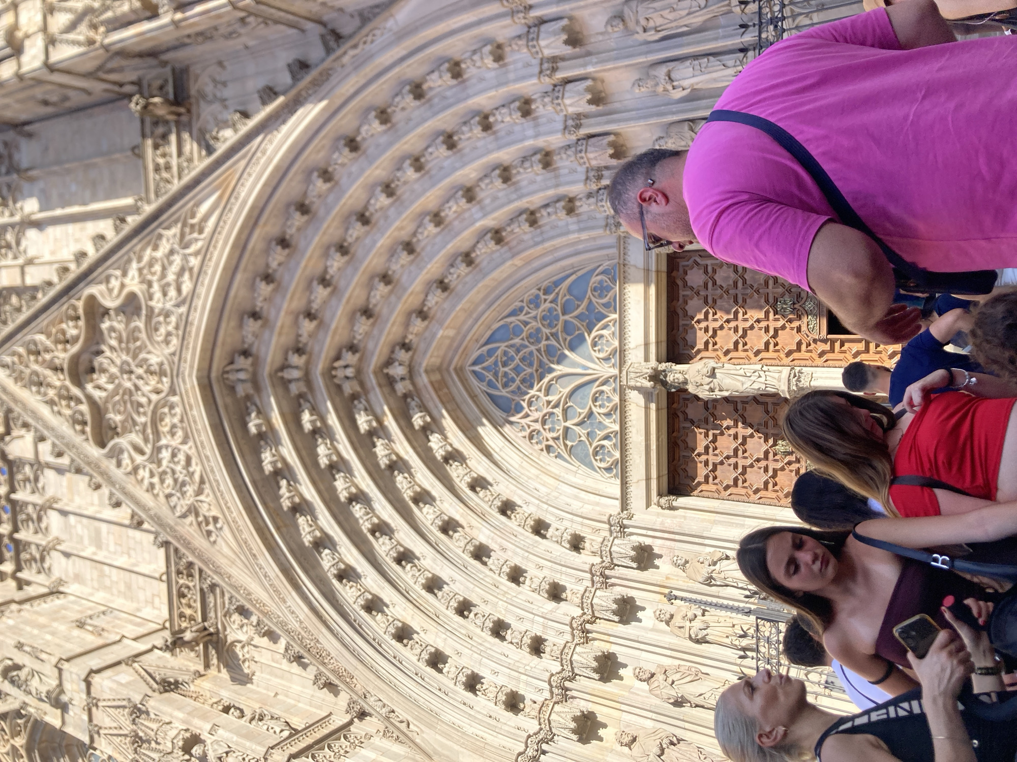 This media file depicts a group of people gathered in front of an ornate architectural facade, likely a cathedral or a historic building in Barcelona, Spain. The architecture features intricate carvings, multiple arches, and decorative stone elements, suggesting a Gothic or Baroque style. Sunlight streams down, indicating it is daytime, likely midday given the brightness and distinct shadows.
In the foreground, several individuals are visible. One person, wearing a bright pink t-shirt and a dark strap across their shoulder, is looking down and to the side, perhaps at a phone or another person. Another person, partially visible in a red top, is also looking downwards. A young woman with long brown hair is looking down and holding a smartphone in her hand. Further down, a woman with blonde hair is also looking down. The people appear to be tourists or visitors, engaged in casual interaction or observing the surroundings.
The overall impression is a bustling tourist spot on a sunny day, with the grandeur of the architecture serving as a backdrop to the everyday activities of the people. The scene evokes a sense of awe at the historical site mixed with the mundane actions of modern life. The orientation of the image is slightly tilted, adding a dynamic perspective. No discernible text is clearly visible in the media file.