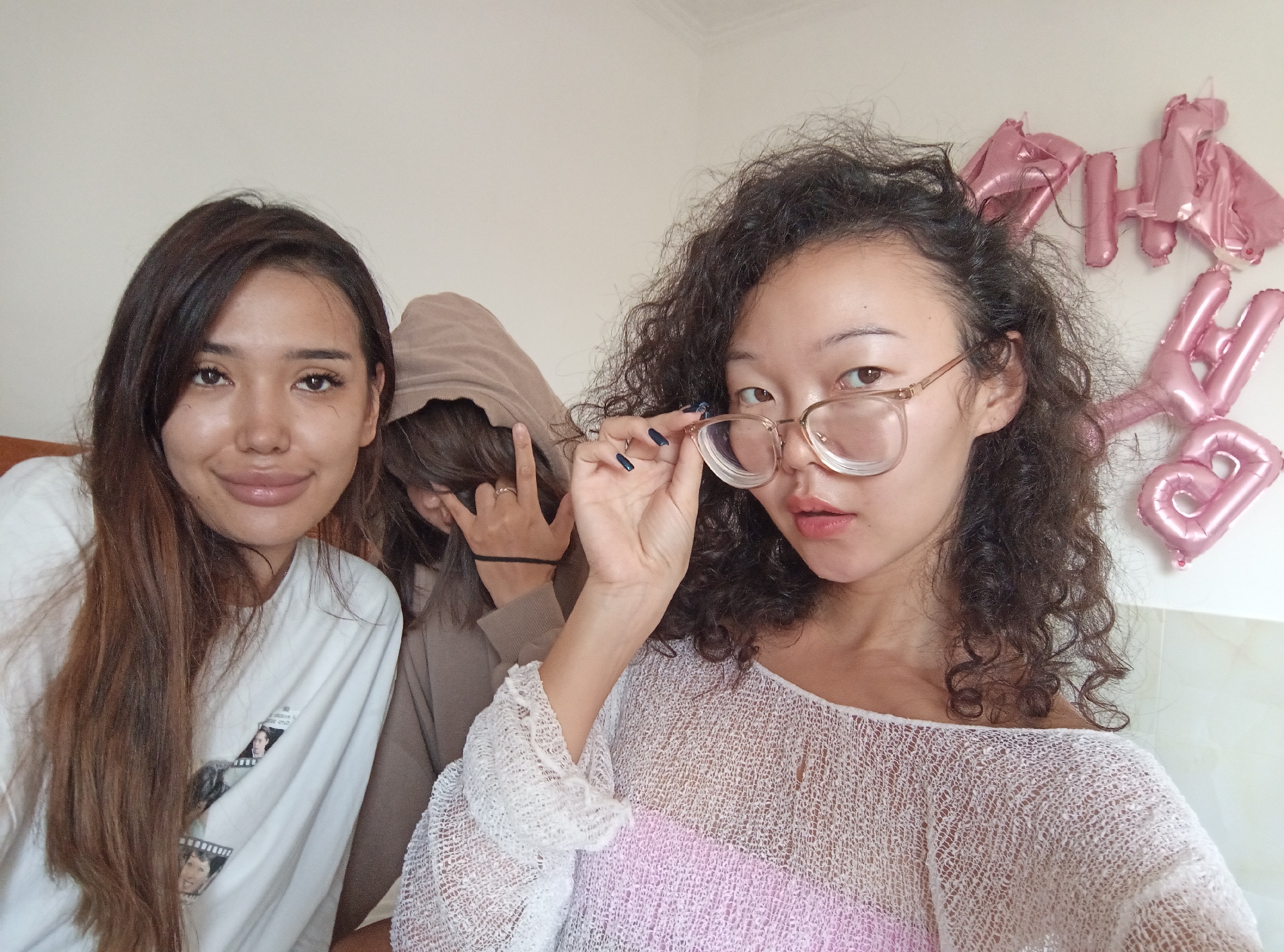 The image shows three young women indoors, likely at a residence in North Kuta, Indonesia. The woman on the left has long brown hair, is wearing a white t-shirt with images, and is facing the camera with a smile. The second woman is wearing a brown hoodie, with the hood pulled up, partially obscuring her face, and she is displaying a hand gesture. The third woman on the right has curly dark hair, is wearing glasses which she is touching, and a white mesh top over a pink tank top. Pink balloon letters spelling "HAPPY BIRTHDAY" are visible on the wall behind her. The background is a plain white wall.