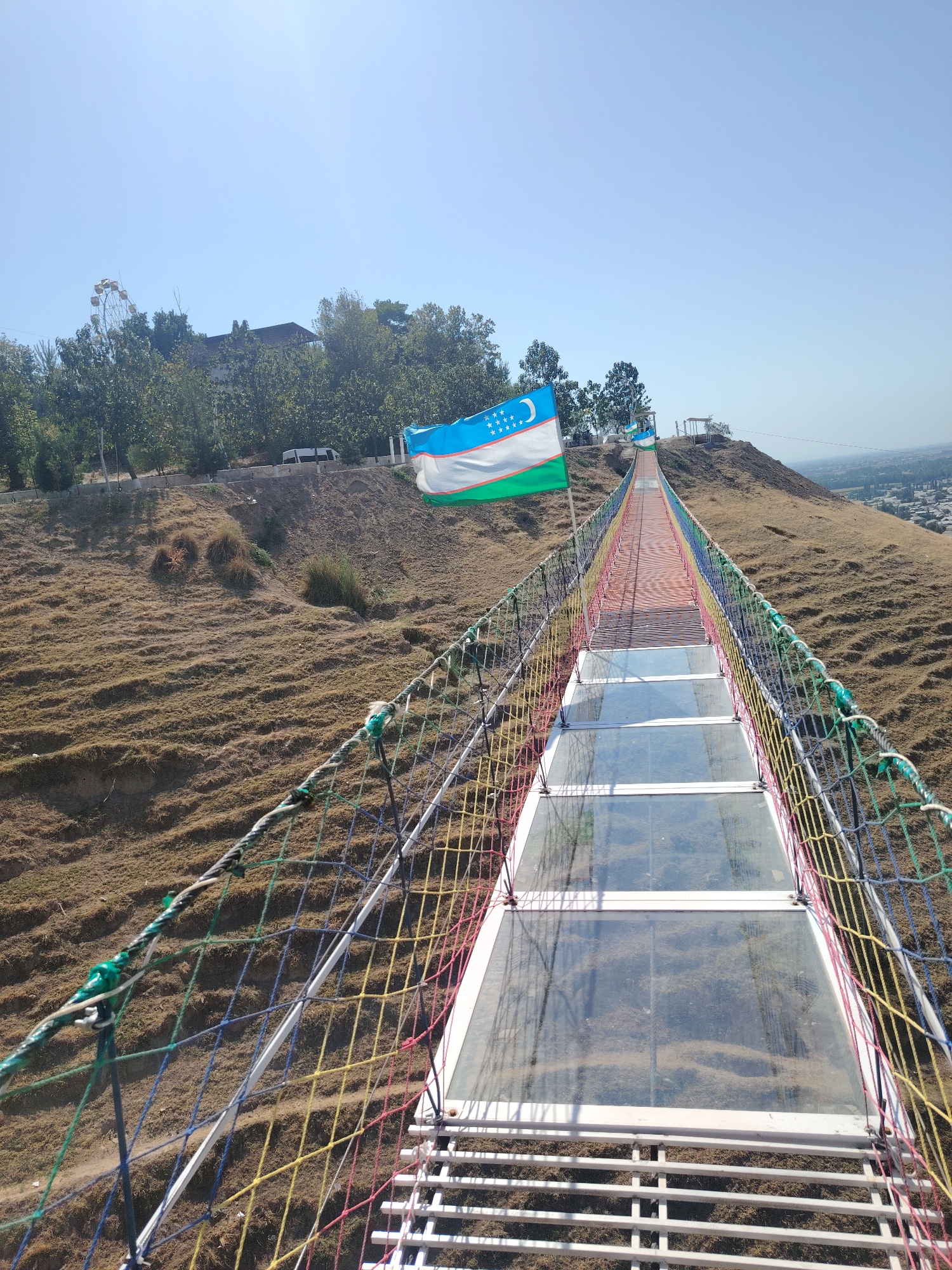 The image shows a view from a suspension bridge with a glass walkway in Xonobod City, Uzbekistan. The bridge is adorned with a colorful rope railing in red, yellow, blue, and green. In the foreground, the walkway is made of transparent glass panels, allowing a view of the dry, grassy terrain below. Two flags of Uzbekistan are visible, one larger and closer to the camera, and another smaller one further down the bridge. The bridge stretches towards a hillside covered with dry grass and scattered trees, with some buildings and a Ferris wheel visible in the distance, suggesting a park or recreational area. The sky is clear and blue, indicating a sunny day. The overall scene depicts a tourist attraction offering panoramic views of the surrounding landscape.