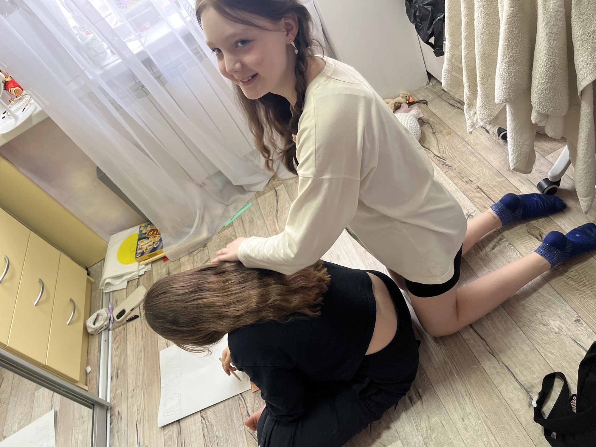 Two young women are on a wooden floor. One woman is kneeling and leaning over the other, who is seated and writing on a piece of paper on the floor. The kneeling woman has on a long-sleeved white shirt, black shorts, and blue socks. The seated woman has brown hair and is wearing black clothing. There is a window with white curtains, a dresser, towels, a black bag, a box, and other items in the background. This location is in the city of Chaikovsky, Russia.