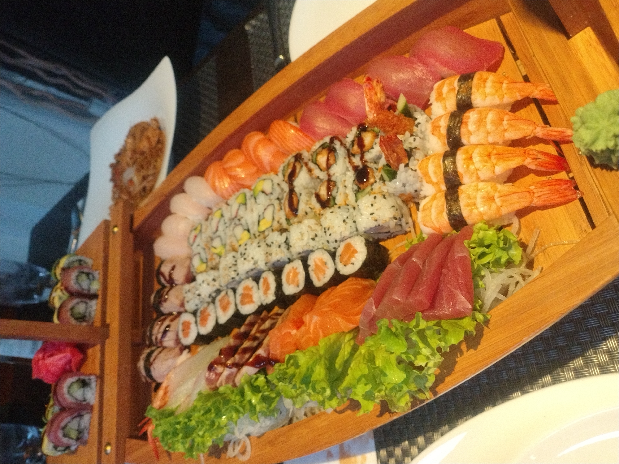 The image shows a large wooden sushi boat platter filled with a variety of sushi and sashimi. Different types of sushi rolls are present, including California rolls, rolls with tuna or salmon, and rolls topped with sauce. Salmon and tuna sashimi are also included. Shrimp nigiri is arranged on the right side of the platter. Green lettuce and seaweed salad are used as garnish. The platter appears to be on a table in a restaurant. There is a small wooden sushi display tower on the left, filled with rolls. A white plate with a crispy, fried appetizer is on the left. This appears to be in a restaurant in Luxembourg.