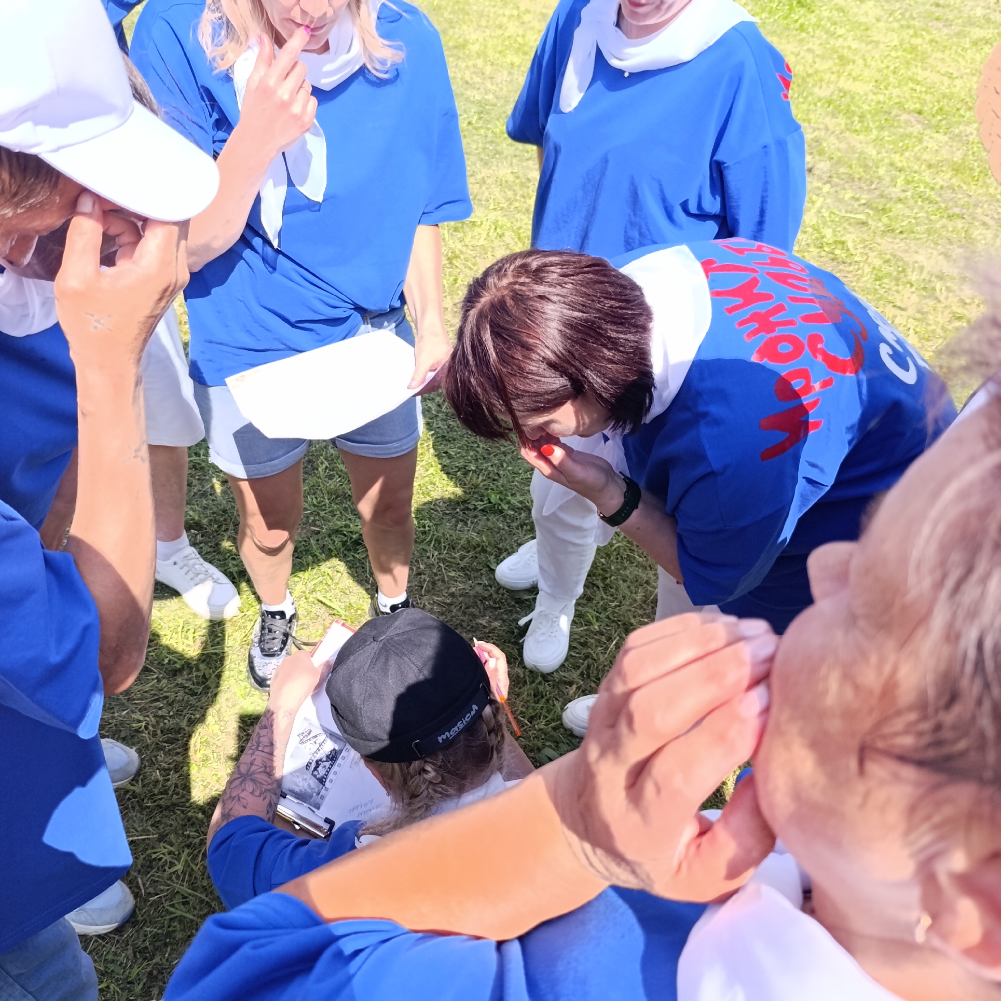 In Prikamskii, Russia, a group of people, some wearing blue shirts and white scarves, are gathered on a grassy outdoor area under daylight. One person, viewed from behind and wearing a black cap, is seated and drawing on a clipboard with a pen. Another individual, also in a blue shirt, is leaning over and appears to be interacting with the person drawing, possibly pointing at the drawing or offering advice. A woman in a blue shirt and white scarf is looking down at her hand, which is near her face. Her hand is raised, and her finger is touching her nose. Another person, wearing a white cap, is partially visible and seems to be looking down at the scene. The ground is covered in short green grass, indicating an outdoor, possibly park-like setting. The clothing of the individuals suggests they might be part of a team or an organized group.