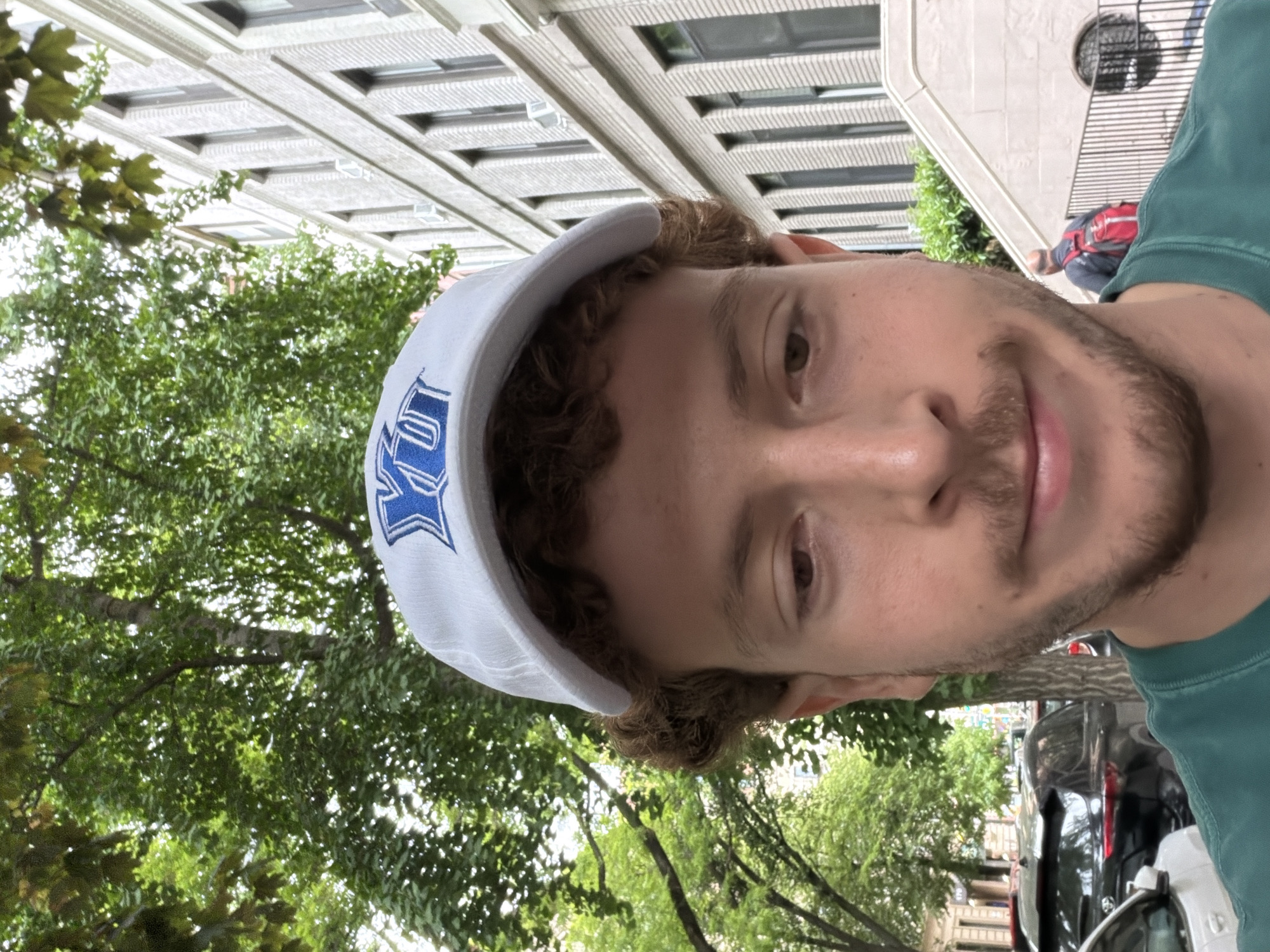 A man is seen from the chest up, with a leafy tree and an apartment building behind him. He is wearing a white baseball cap with a blue logo. The photo was taken outside in daylight in a city that is likely to be New York, United States of America. A car can be seen parked in the background.