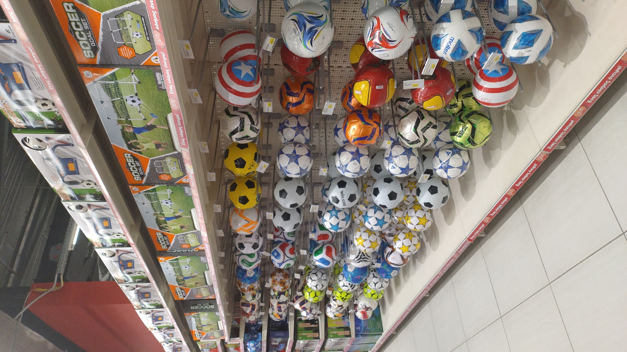 The image depicts a retail display of soccer balls and soccer goal sets in what appears to be a store. The shelves are stacked with various soccer balls of different designs, colors, and sizes. Some of the soccer balls are branded with logos. Boxes of "Soccer Goal Set" are also visible on higher shelves, featuring illustrations of children playing soccer. The scene is well-lit, suggesting an indoor commercial environment. The retail setting is consistent with a toy store or sporting goods store. The floor is tiled with a light-colored material. Based on the signage, which includes Greek text "Ένας κόσμος όνειρο!" (meaning "A world of dreams!"), the location is likely a store in Greece. Specifically, based on the store's signage and product offerings, this image was taken at a Jumbo store in Pylaia Municipal Unit, Greece, which is located in the Thessaloniki regional unit.