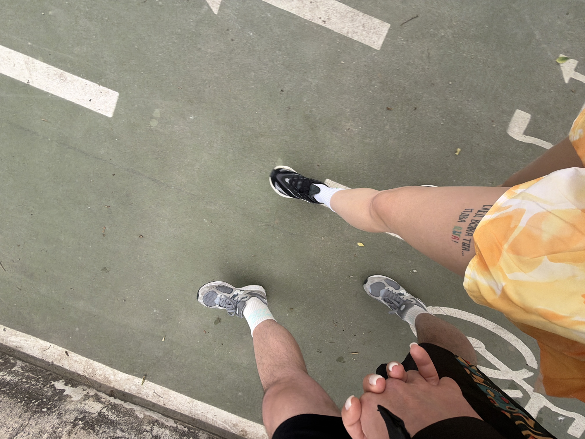 A high-angle shot depicts two people holding hands, standing on a paved surface marked with white arrows and a circular shape, potentially indicating a bike lane or pedestrian path. The location is assumed to be Arroyo de la Miel-Benalmádena Costa, Spain.
One person wears black running shoes with white socks and black shorts. The other person wears black and white running shoes with white socks, a yellow floral patterned garment, and has a tattoo on their upper thigh.
The surrounding environment includes a concrete curb or edge visible on the left side of the image, suggesting a sidewalk or designated path within the urban area. The overall scene implies a casual outdoor setting, likely a public space used for recreation or transportation.