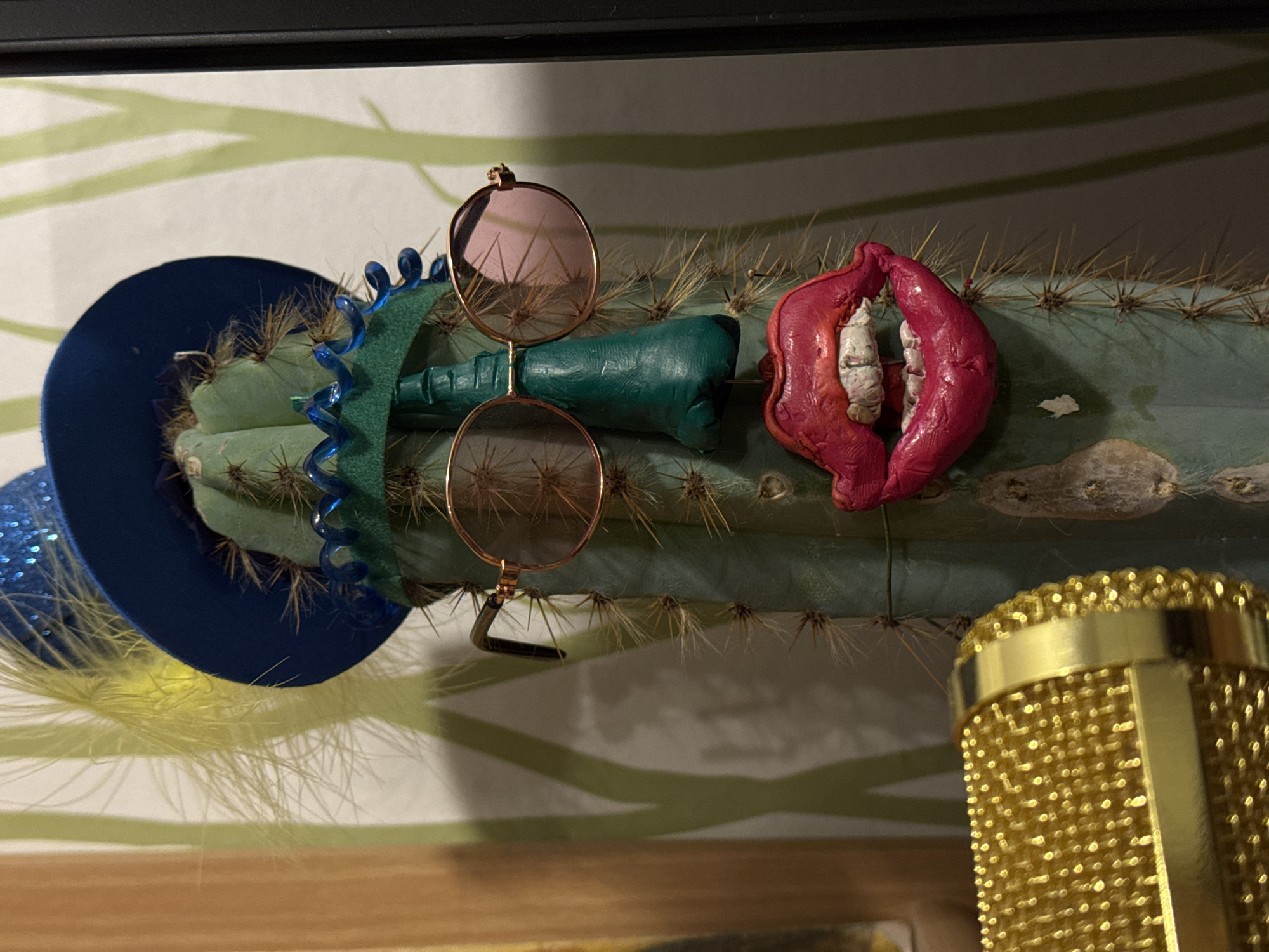 The image displays a whimsical and surreal indoor still life, centered around a pale green columnar cactus lying horizontally. The cactus is adorned with an array of humorous accessories, creating an anthropomorphic character.
On the left side of the cactus, a miniature dark blue sombrero is perched, featuring glitter on its brim and a fluffy yellow feather protruding from underneath. A bright blue spiral ornament is also attached near the hat. In the middle section of the cactus, a pair of round, rose-tinted sunglasses with delicate gold frames rests. Directly beneath the sunglasses and extending to their right are two small, dark green, textured objects, resembling stylized bottles or decorative elements. Further to the right, a striking bright pink object, shaped like exaggerated, open lips with rough, white tooth-like structures inside, is impaled onto the cactus spines. The cactus itself has prominent ribs and numerous sharp, light-colored spines.
In the bottom right corner of the frame, a golden microphone with a distinct mesh texture is partially visible, suggesting it's part of the surrounding environment or another artistic arrangement. The background features a light-colored surface, likely a wall, decorated with a subtle, abstract pattern of pale green, wave-like lines. A wooden surface is visible at the very bottom edge of the image.
No people are present, and no specific activity or event is taking place; the scene is a static arrangement. The lighting suggests an indoor setting, possibly under artificial light, without any indications of outdoor weather or specific time of day. The overall impression is one of playful creativity. There is no visible text in the image. The provided location context for this media file is Ново-Никольское, Russia.