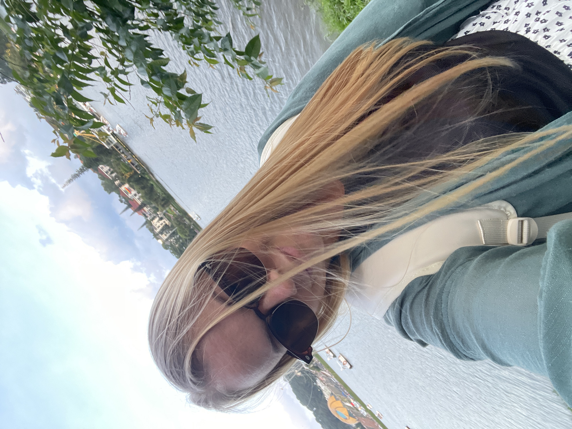 A female individual with long, blonde hair and dark sunglasses is depicted in a selfie, angled slightly downward. She wears a teal-colored cardigan over a black top, and a portion of a white skirt with a dark pattern is visible. A light-colored backpack strap is present over her right shoulder. The background features a large body of water, identified as Xuan Huong Lake in Da Lat, Vietnam, with ripples on its surface. Multiple small boats are scattered across the lake. On the opposite shore, various buildings, including some with spired roofs and red tiles, are situated on a hillside. A large, colorful structure, resembling a deflated hot air balloon or similar exhibition, is visible on the left side of the lake. Green foliage and grass line the immediate foreground on the right. The sky overhead is light blue with scattered white and grey clouds, indicating a daytime setting.