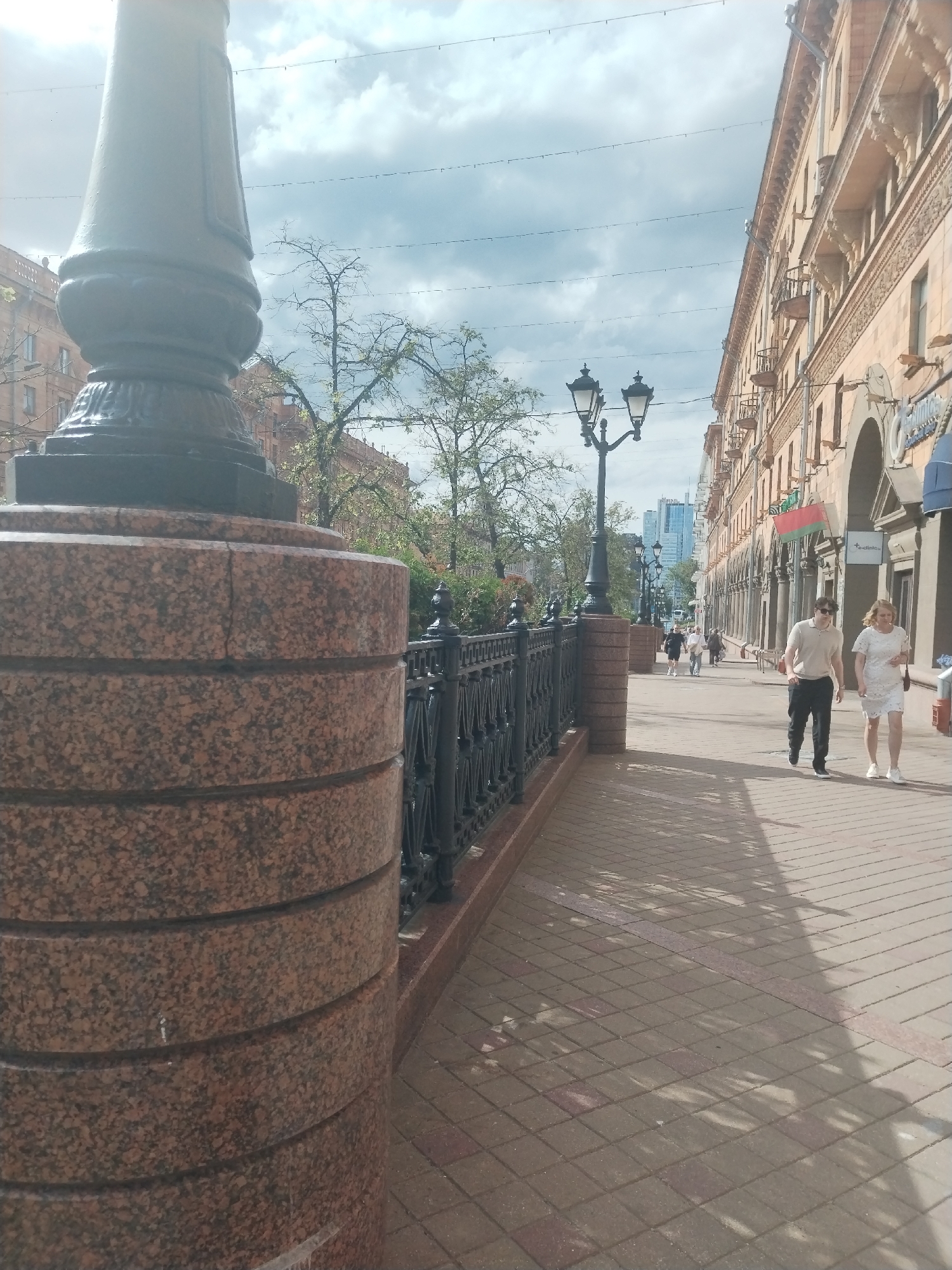 The image captures a daytime street scene in Minsk, Belarus, likely on a central thoroughfare. The perspective is from a low angle, looking down a wide paved sidewalk bordered by a decorative fence and a multi-story building.
In the immediate foreground on the left, a large, cylindrical lamppost base constructed from reddish-brown granite with dark speckles is prominent. It is composed of stacked, rounded sections. To its right, and extending into the midground, is a black wrought-iron fence featuring ornate scrollwork, mounted on a matching granite base.
The sidewalk, covering the lower right quadrant of the image, is paved with light-colored (beige/grey) square pavers interspersed with some reddish-brown rectangular pavers, laid in a diagonal pattern. Sunlight casts distinct, elongated shadows across the sidewalk from objects off-frame to the left, likely trees or additional lampposts.
Along the right side of the street, a long, multi-story building with a light-colored, ornate facade dominates the view. Its ground floor features a series of arched openings, some housing storefronts. The upper floors display balconies and intricate architectural detailing, including pilasters and patterned friezes. A Belarusian national flag (red horizontal stripe over a green horizontal stripe, with a vertical white ornamental pattern on the hoist side) is prominently displayed on the building's facade. A partial sign for a business, possibly reading "teclinka," is visible on one of the ground-floor storefronts.
Several pedestrians are walking on the sidewalk. Closest to the viewer, a man and a woman are walking side-by-side towards the camera. The man, on the left, wears a light-colored polo shirt, dark trousers, and dark shoes, with sunglasses. The woman, on the right, wears a white, short-sleeved lace or patterned dress and white sneakers, carrying a dark shoulder bag. Further down the street, additional pedestrians are visible, some walking towards, others away from the viewer.
In the midground and background on the left, behind the fence, are trees with varying degrees of foliage, some appearing sparsely leaved. Another ornate black lamppost with dual lights is visible further down the street on the left. In the far distance, towering modern high-rise buildings are discernible against the sky, indicating a city skyline. The sky above is partly cloudy, with areas of bright light suggesting sunlight breaking through.