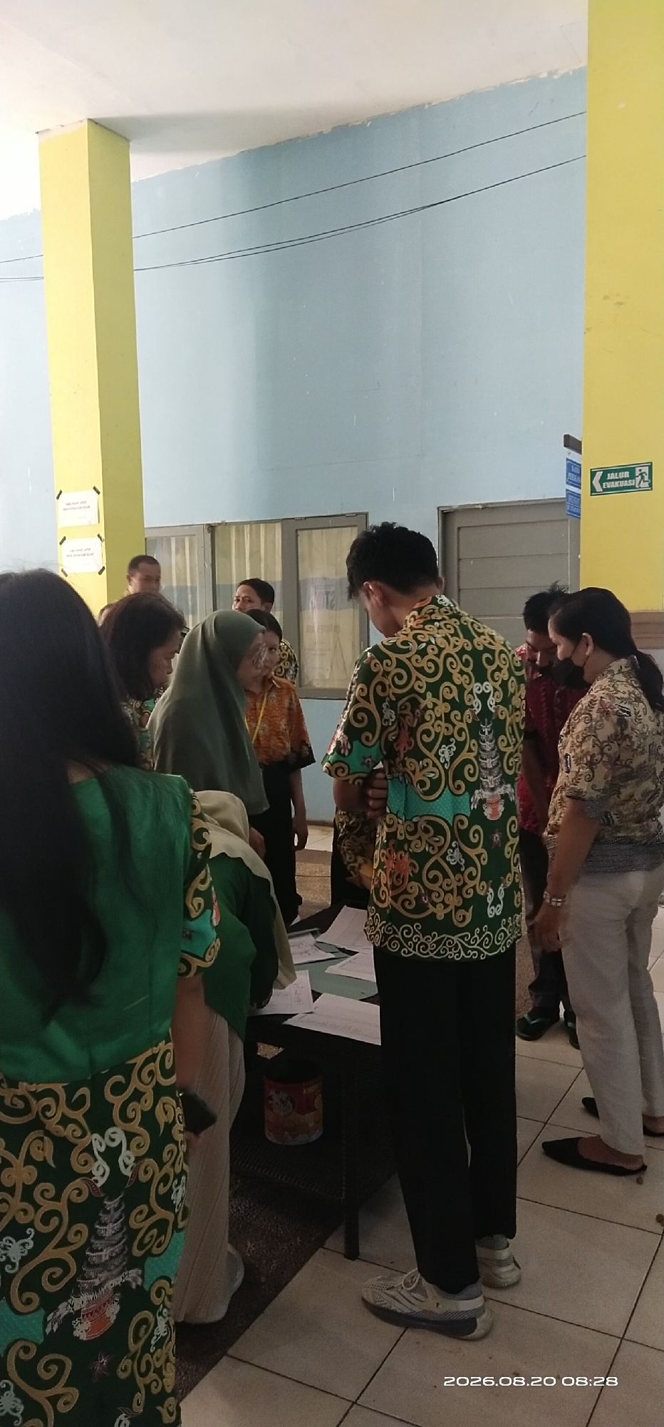 The image shows a group of people standing around a table with papers on it. The people are wearing batik shirts and headscarves, suggesting a formal or cultural event. One person is wearing a green batik shirt with a repeating pattern and intricate designs, including a pagoda-like structure. Another person is wearing a green headscarf and a similar green batik shirt. A man in a green batik shirt with a similar pattern is also visible. The environment appears to be indoors, possibly a community center or an office building, with light blue walls and a yellow pillar on the left side of the frame. There is a sign with text and an arrow on the yellow pillar, indicating an evacuation route. The floor is tiled with a light brown and white pattern. The overall scene suggests a gathering or a meeting taking place in Tamiang Layang, Indonesia, given the Indonesian clothing and the presence of a sign with "JALUR EVAKUASI" (Evacuation Route) which is common in public buildings in Indonesia. The date stamp at the bottom right corner indicates "2026.08.20 08:28".