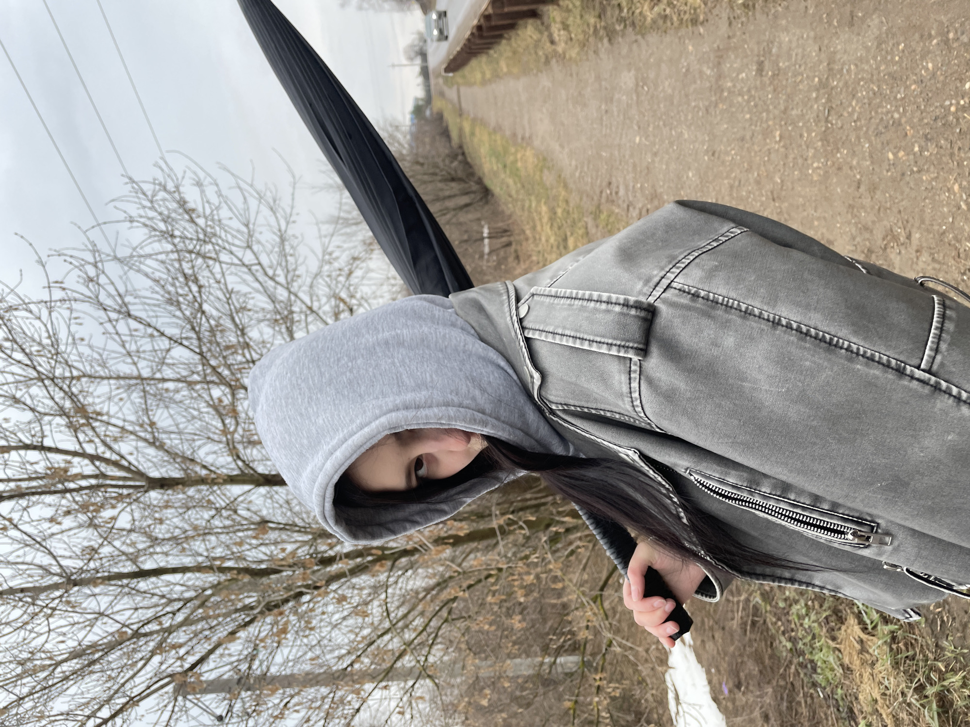 A woman in a gray hooded sweatshirt and gray jacket is walking along a dirt path next to a snow-covered field. She is holding a black umbrella. The sky is overcast. In the distance, there is a road with a car. The location is near Фоки, Russia.