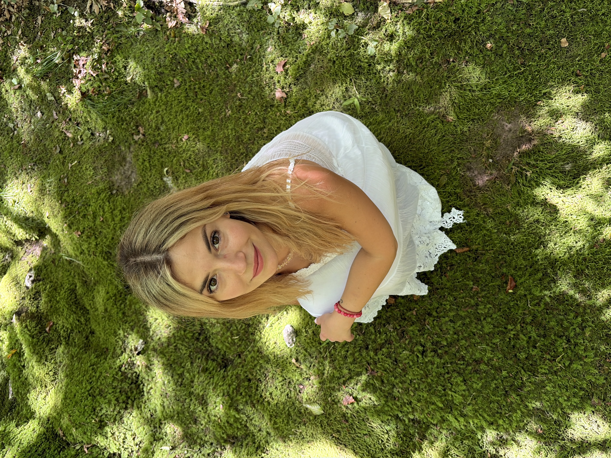 A young woman with blonde hair is crouched on a moss-covered ground, looking up at the camera. She is wearing a white dress with lace detailing and a pink bracelet on her left wrist. The ground is covered in lush green moss, with some fallen leaves scattered around. Sunlight filters through the trees, creating dappled shadows on the ground. The scene appears to be in a natural outdoor setting, possibly a forest or park. The location is Vusanje, Montenegro.