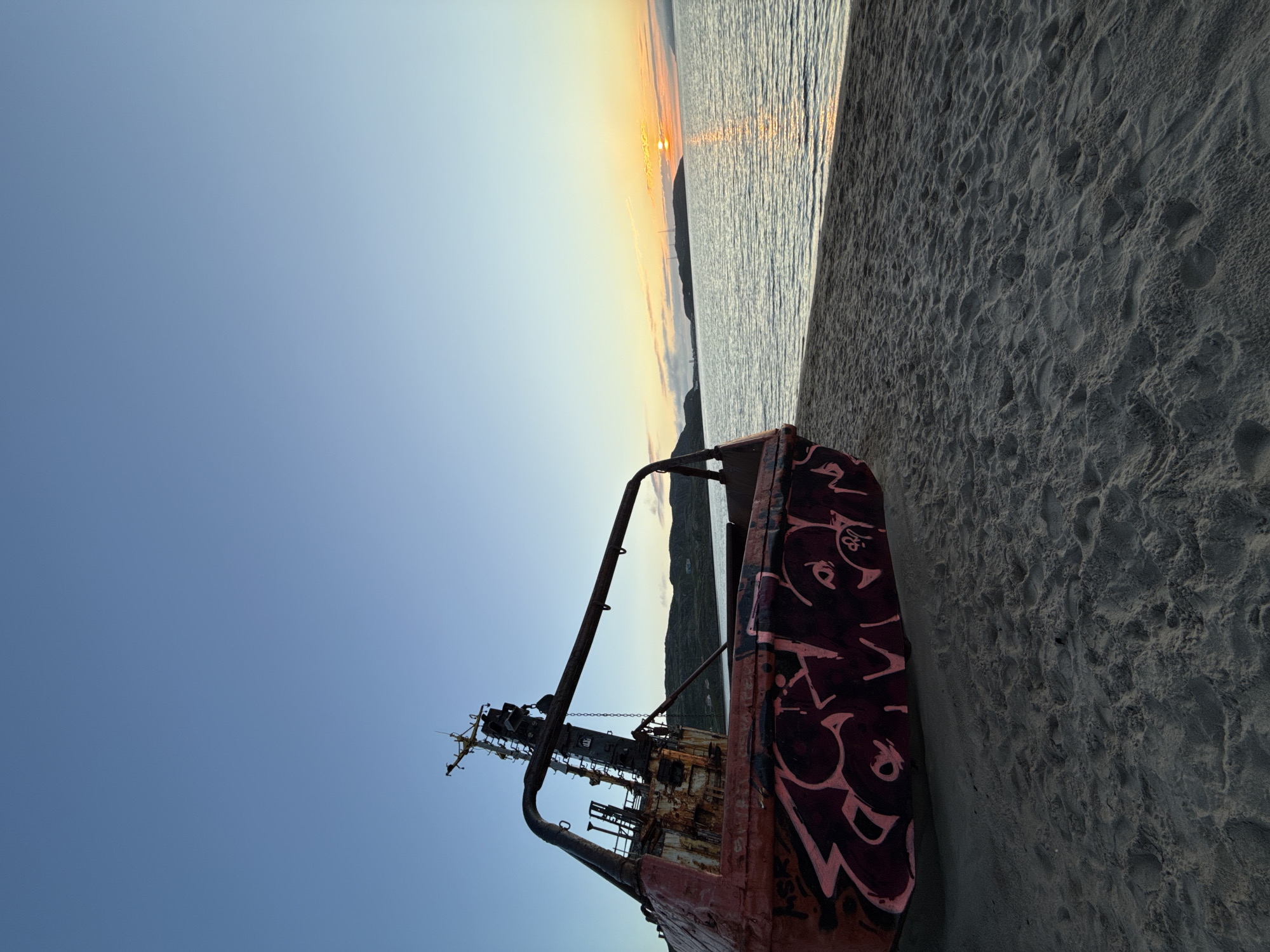 This image captures a sunset scene on a sandy beach in Teriberka, Russia. In the foreground, the rusted hull of a beached ship dominates the left side of the frame. The ship is covered in graffiti. The sand on the beach is textured with footprints. The ocean is visible in the mid-ground, with gentle waves reflecting the warm hues of the setting sun. The horizon is marked by a chain of hills and a faint outline of a town or structures. The sky transitions from a clear, pale blue at the top to a soft gradient of yellow and orange near the horizon, where the sun is setting. The overall atmosphere is serene and somewhat melancholic.