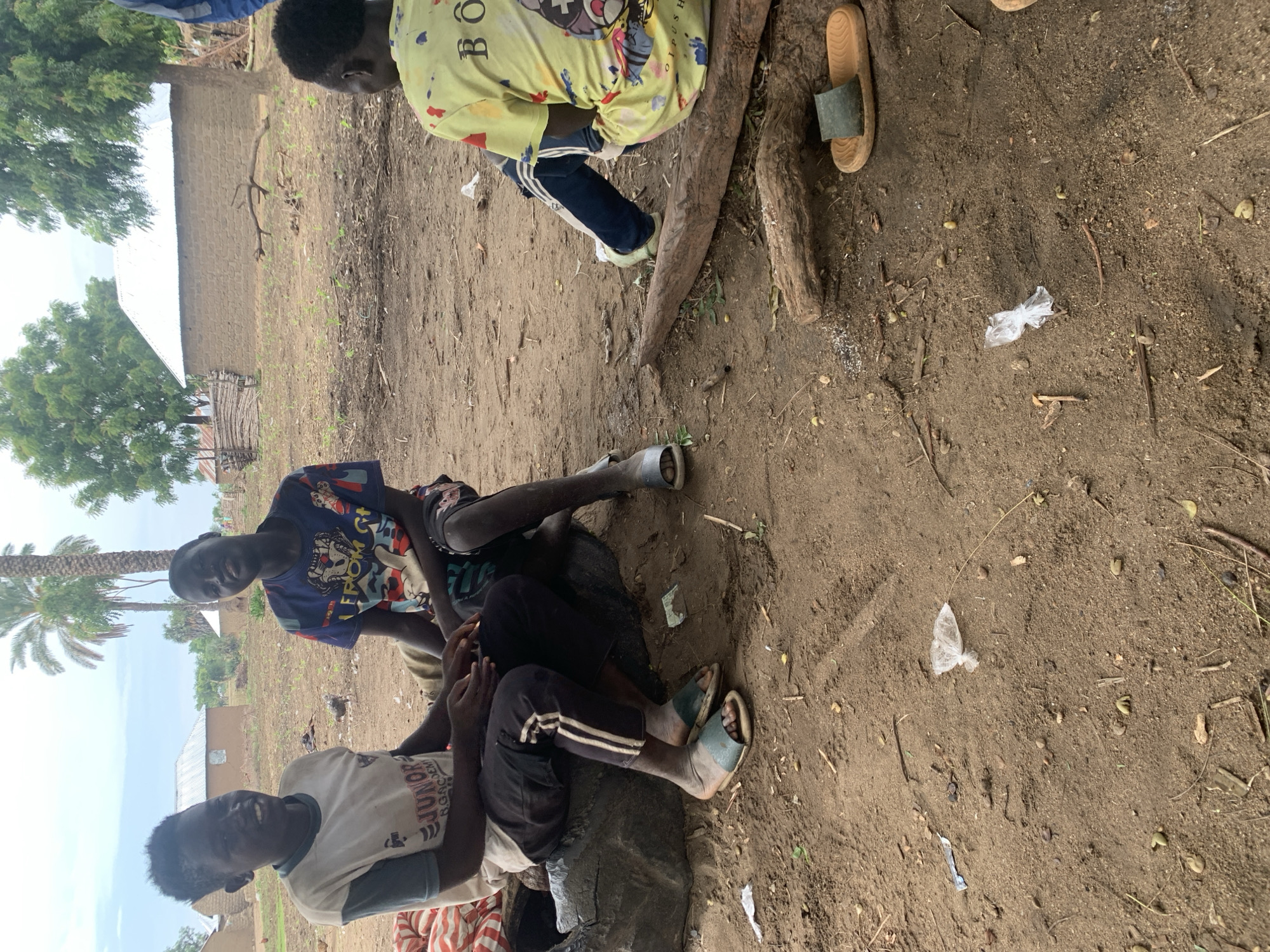 This image depicts a group of young people sitting on the ground in what appears to be an outdoor, possibly rural, setting in Dull A, Nigeria. The ground is a mixture of dirt and sand, with scattered leaves, twigs, and some debris, including small pieces of plastic. In the background, there is a building with a corrugated metal roof and a brick wall. Lush green trees are visible on the left side of the image, suggesting a somewhat natural environment.
There are at least four individuals visible, though their full bodies are not always in frame. Two of the individuals are more prominently featured in the foreground, appearing to be conversing or interacting. One person is wearing a light-colored t-shirt with a graphic design and the text "JUNIOR RGACMEN" partially visible. Another individual is wearing a dark blue t-shirt with a colorful graphic. A third person is seated behind them, wearing a yellow t-shirt with a graphic and text that includes "BO" and possibly other obscured letters. The fourth person's clothing is mostly out of frame, but they are wearing dark pants and a t-shirt.
The individuals are dressed casually, and their postures suggest a relaxed atmosphere. One person is wearing blue and white striped pants and light blue sandals, while another is wearing dark pants with white stripes and light blue sandals. A third person has a brown sandal to their right, and another dark sandal is visible near the foreground.
The lighting suggests it is daytime, possibly mid-morning or afternoon, with diffused light and no strong shadows, indicating overcast weather or a shaded area. The overall scene conveys a sense of everyday life and social interaction among young people in a communal outdoor space.