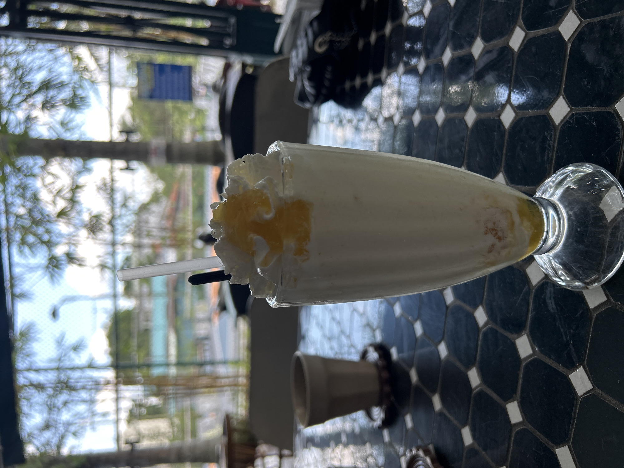 A milkshake with whipped cream and a yellow topping sits on a black and white tiled table. Two straws, one clear and one black, are in the milkshake. In the background, out of focus, is a view of a street with trees and buildings through a window. A small, tan ceramic cup sits on a coaster to the left of the milkshake. The image appears to be taken outdoors or in a location with a large window offering a view of the outside. There is no visible information to confirm the location in Thanh Hưng, Vietnam.