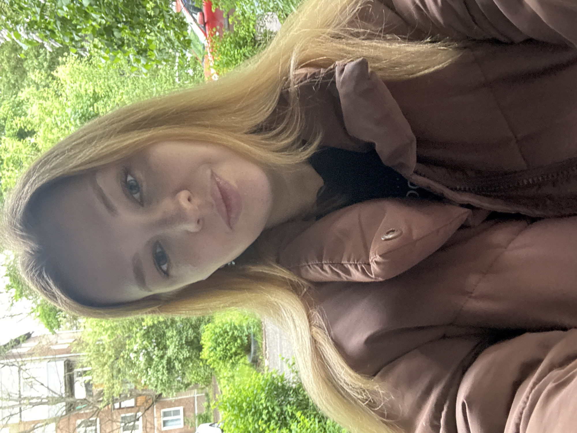 A woman with long blonde hair and fair skin wears a brown puffer jacket. She is outdoors in an area with green foliage, including trees and bushes. In the background, there are buildings that appear to be residential, and they may be in the city of Заволжье, Russia, although the location is not definitively confirmed. She appears to be taking a selfie.