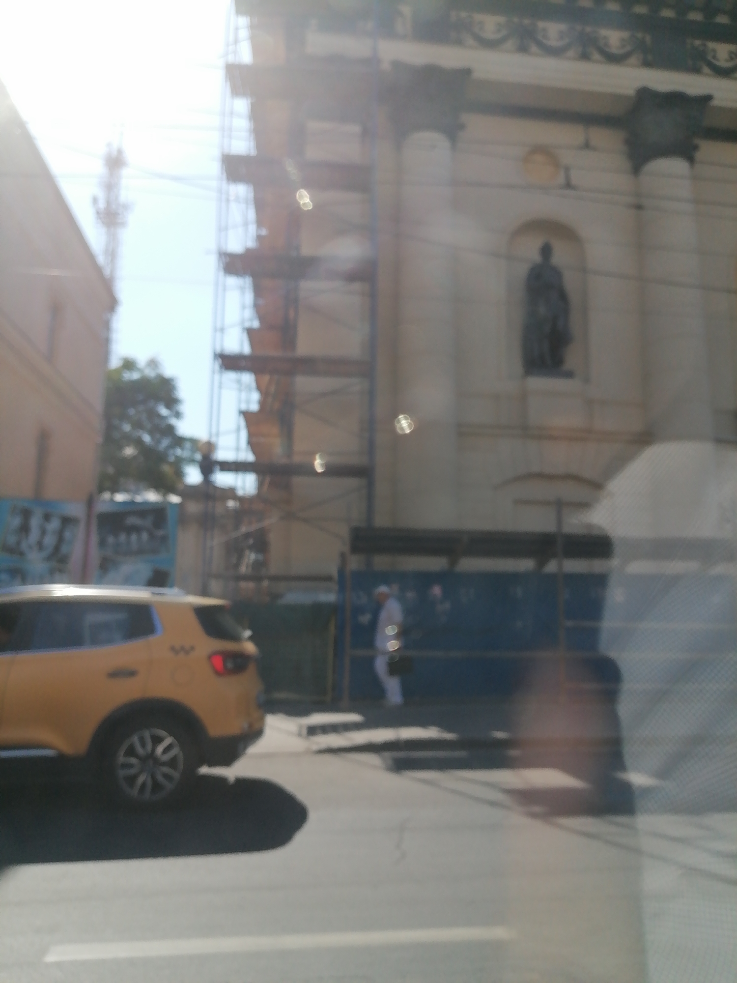 The image depicts a street scene in Rostov-on-Don, Russia, under bright, sunny conditions that create significant lens flare and glare. In the foreground, a yellow car is parked or moving slowly. To the left, part of a building is visible, appearing to be under construction or renovation, with scaffolding extending upwards. Behind the scaffolding, a tall structure, possibly a radio tower, is faintly discernible against the sky. In the mid-ground, a person dressed in white is walking along a sidewalk, partially obscured by a blue barrier or fence. Behind this, a large building with classical architectural elements, including columns and a statue in an alcove, is visible. The overall impression is of a bustling urban environment with ongoing construction work.