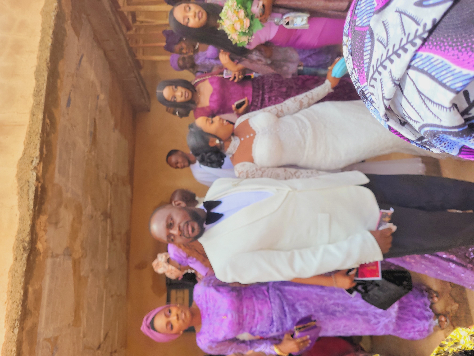 The image captures an indoor or covered outdoor gathering in Bukuru, Nigeria, featuring multiple individuals. The ceiling is rough, light brown concrete with visible structural elements. In the foreground, a dark-skinned adult male stands slightly left of center, wearing a white tuxedo jacket with a black lapel, a white vest, a white shirt, and a black bow tie, with dark trousers. He holds a small black bag and a mobile phone in his left hand, looking towards the right. To his right, a dark-skinned adult female, presumed to be a bride, wears a white, long-sleeved, high-neck wedding gown with lace details and a necklace, smiling and looking downwards. She holds a light blue object in her right hand.
To the left of the central male, a dark-skinned adult female wears a purple traditional outfit and a matching head wrap, holding a purple clutch bag. Behind the central female, another dark-skinned adult female wears a dark purple top and matching skirt, holding a mobile phone and a dark object, looking forward. To the far right, a dark-skinned adult female in a pink dress holds a bouquet of light pink and cream flowers with green foliage. Several other individuals, primarily women in various purple attires and head wraps, and at least one male in white traditional wear, are visible in the blurred background, observing the scene. A multi-colored fabric with purple, blue, and white patterns is partially visible in the lower right foreground.