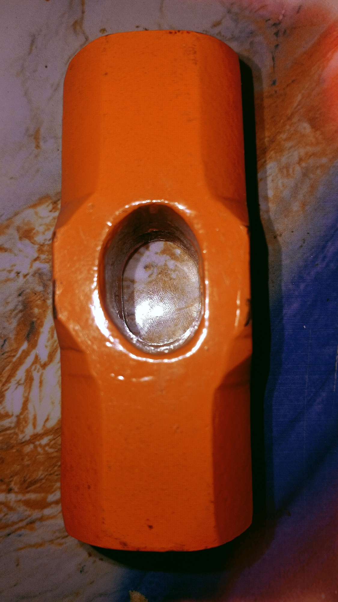 An orange sledgehammer head, with an oval-shaped opening for a handle, rests horizontally on a patterned surface that appears to be a countertop or table. The surface has a marble-like appearance with swirls of brown, beige, and gray. To the right of the sledgehammer, a blue surface with a grid pattern is visible, suggesting a different material or background. The lighting is harsh, creating strong highlights and shadows, particularly on the metallic interior of the sledgehammer's opening. The image is a close-up shot, focusing entirely on the tool and its immediate surroundings. There are no people or discernable actions taking place. The location in the city of Pematang Danau, Indonesia, is not visually represented in the image.