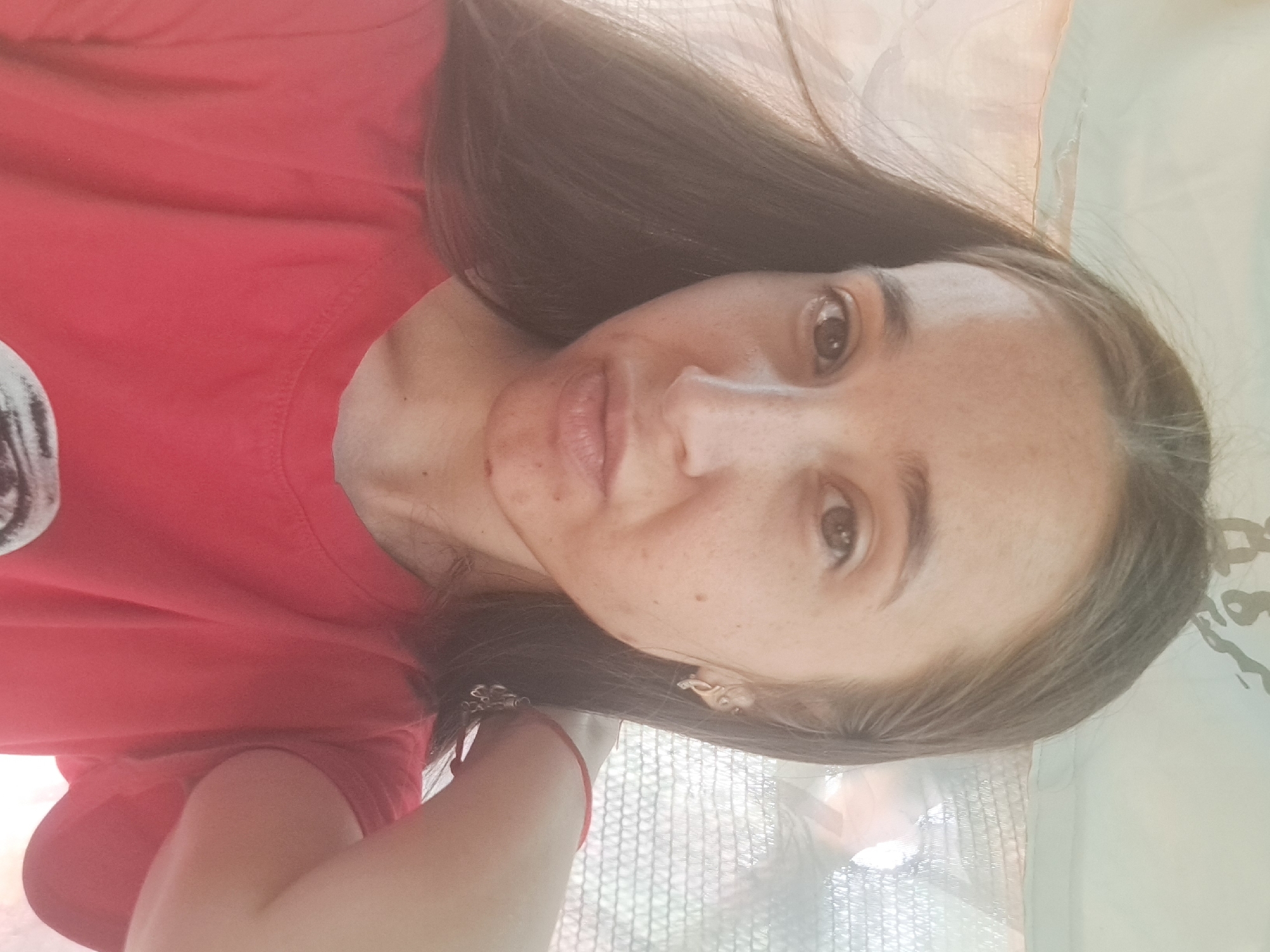 A young woman with long, brown hair and light brown eyes is pictured in a close-up, selfie-style photograph. She has a fair complexion with some freckles and appears to be wearing a red t-shirt. Her hair is slightly windblown, and she is looking directly at the camera. The background is softly blurred and suggests an outdoor setting, possibly a shaded area with a patterned screen or fabric visible. She is wearing an earring in her left ear and a bracelet on her right wrist. The lighting in the image is soft, and the overall tone is warm. There are no identifiable landmarks or specific details that would indicate the location within the city of Vypasne, Ukraine.