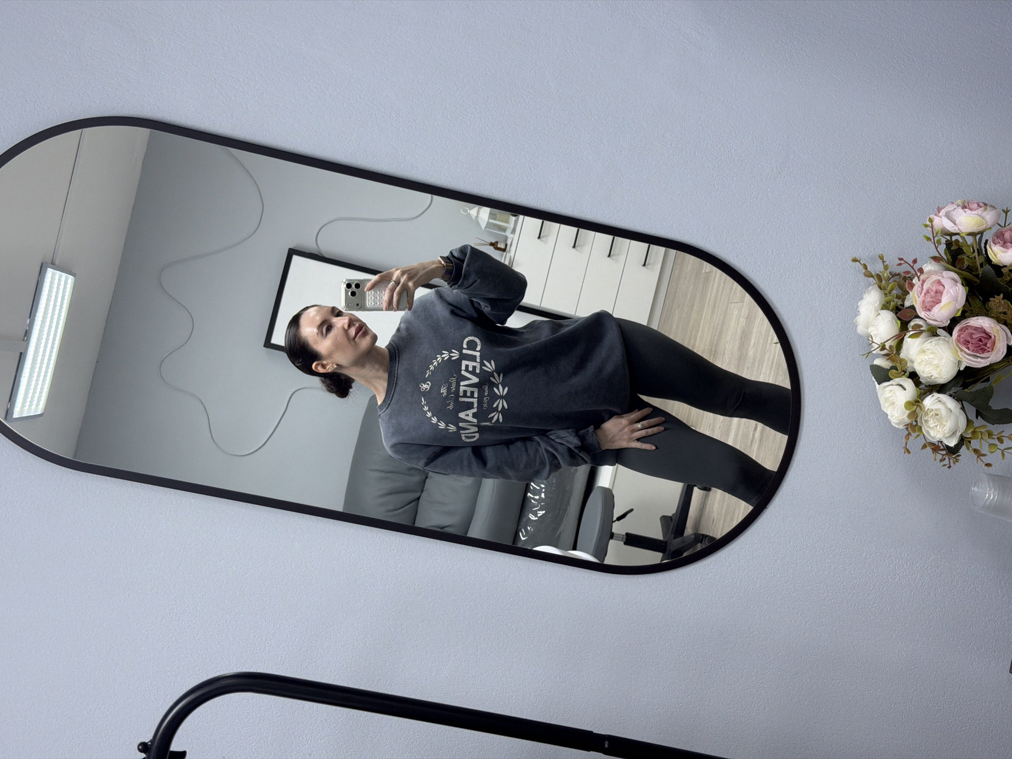 A woman is taking a picture of herself in a mirror. She has dark hair pulled back and is wearing a gray sweatshirt with the word "Cleveland" printed on it in reverse. She is wearing black leggings. The mirror has a black frame and an oval shape. In the mirror's reflection is a gray couch, a white dresser, and a framed print. Next to the mirror is a bouquet of pink and white flowers in a vase. The wall is light blue. A black metal object is partially visible in the bottom left corner of the image. There is no evidence to suggest the photo was taken in Perm, Russia.