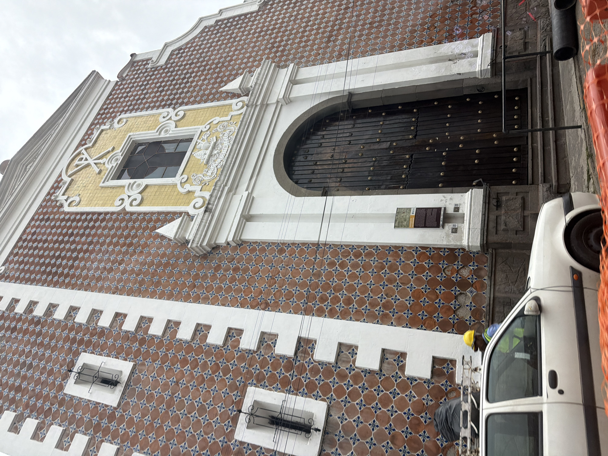 The image depicts a portion of the facade of a building in Puebla, Mexico, characterized by its unique architecture. The building features brown and blue patterned tiles across its surface, punctuated by white architectural details. These details include a horizontal band of tooth-like protrusions and decorative window frames. A large, arched wooden door, studded with metal accents, is prominent on the right side of the image.
A white van is parked in the foreground, partially obscuring the lower right portion of the building. A man wearing a yellow hard hat and high-visibility vest is visible near the van, possibly engaged in some form of work or maintenance. The scene includes utility wires and a section of orange construction netting, further indicating ongoing activity in the vicinity. The sky appears overcast.