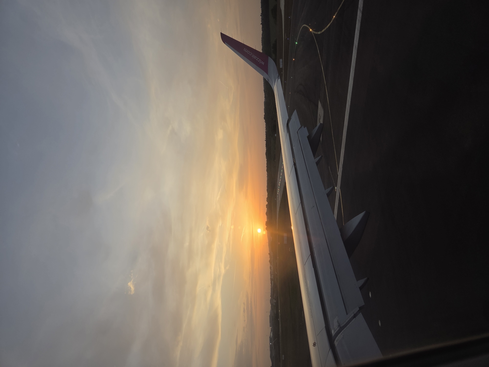 The image shows a view from an airplane window during sunset. Part of the plane's wing is visible, with the website "wizzair.com" printed on the winglet. Below the wing, the ground is visible, suggesting the plane is either taking off or landing. A runway with lights and markings is visible on the ground. The sun is setting on the horizon, casting a golden glow across the sky and land. The location is North Hertfordshire, United Kingdom, as identified by the airport environment and airline details. There are no people directly visible in the image. The focus is on the sunset and the airplane wing.
