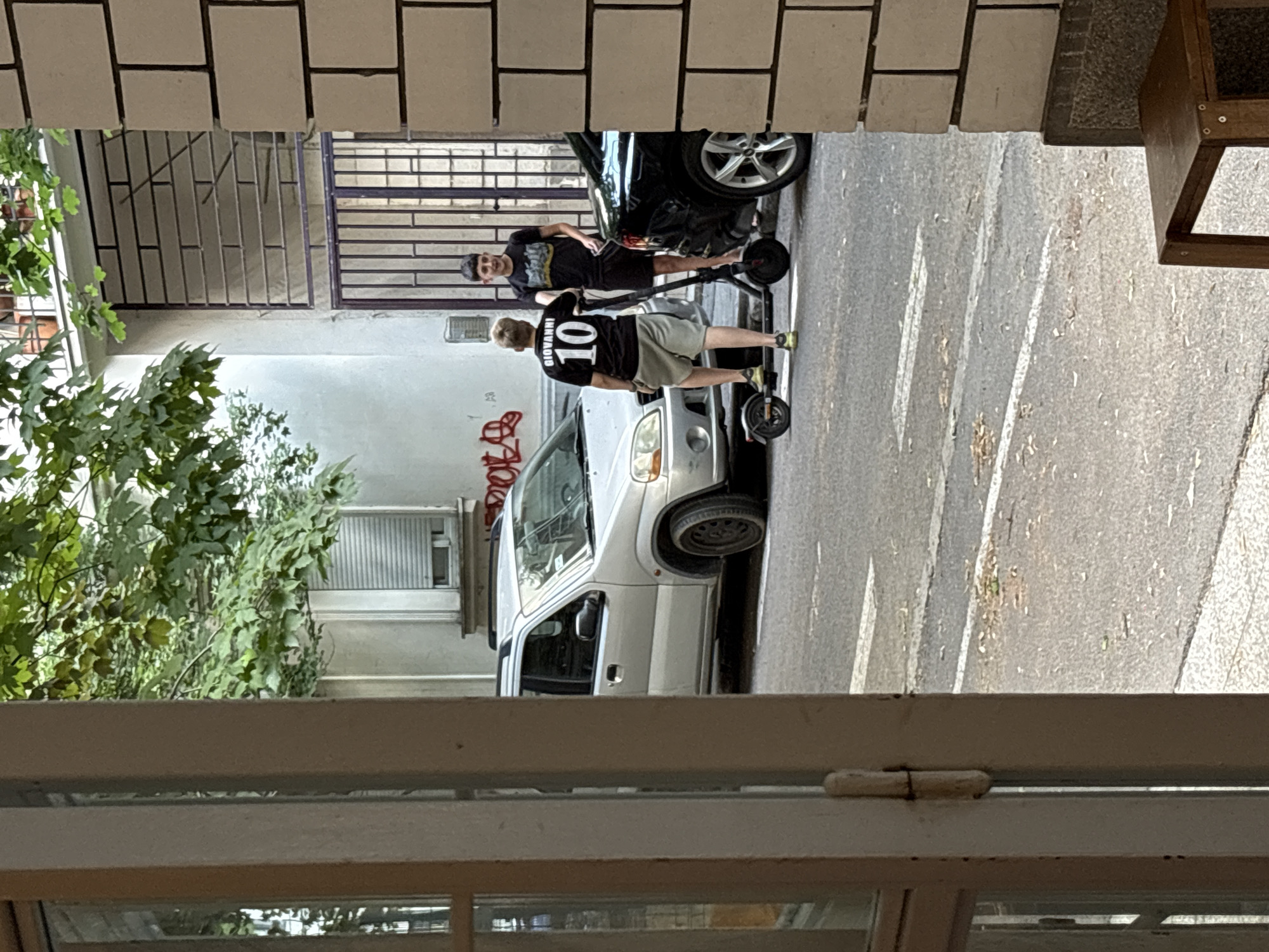 The image captures an outdoor urban scene, likely in Belgrade, Serbia, depicting two young males near parked vehicles and on scooters. In the foreground, a partial view of a doorway or window frame with a hinge suggests an indoor vantage point. The background reveals a narrow street lined with buildings. A silver hatchback is parked on the left side of the street, while a black sedan is parked on the right. Two young men are present. One, facing away from the camera, is on a kick scooter and wears a black t-shirt with "GIOVANNI 10" printed on the back, and light-colored shorts. The second young man, facing the other, stands with a black kick scooter and wears a black t-shirt and shorts. Trees with green foliage overhang the scene, and there is graffiti on the building wall. The ground is asphalt, with visible white road markings. The overall impression is a casual moment in a residential or mixed-use urban environment.