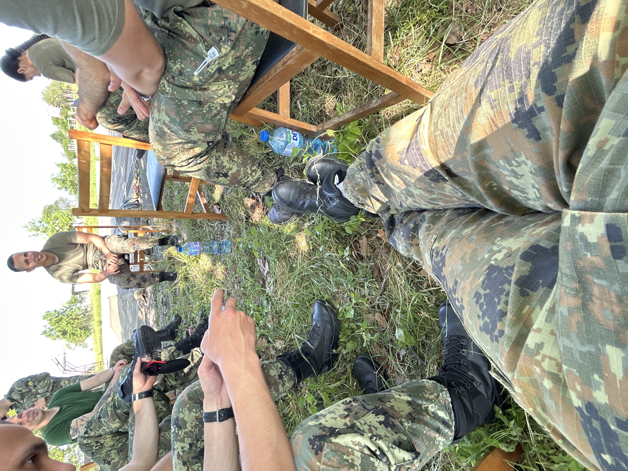 A group of people dressed in military camouflage are seated on folding chairs in a grassy area. The scene is in daylight. Most individuals are partially visible, from the knees up. All are wearing black boots. A blue water bottle sits on the ground near the chairs. The location is Veliko Tarnovo, Bulgaria.