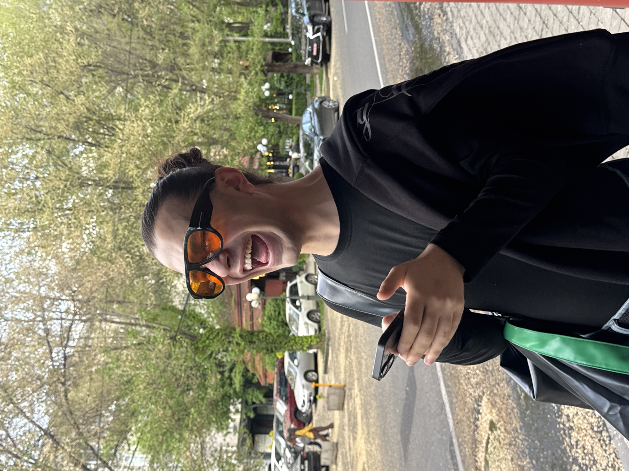 A man in dark clothing and orange-tinted sunglasses is laughing on a sidewalk in Yerevan, Armenia. The man has his hair pulled back and is wearing a black t-shirt, a black jacket, and carrying a black bag with a green strap. He is holding a black phone in his hand. Behind him is a street lined with parked cars and trees. The buildings in the background appear to be residential.