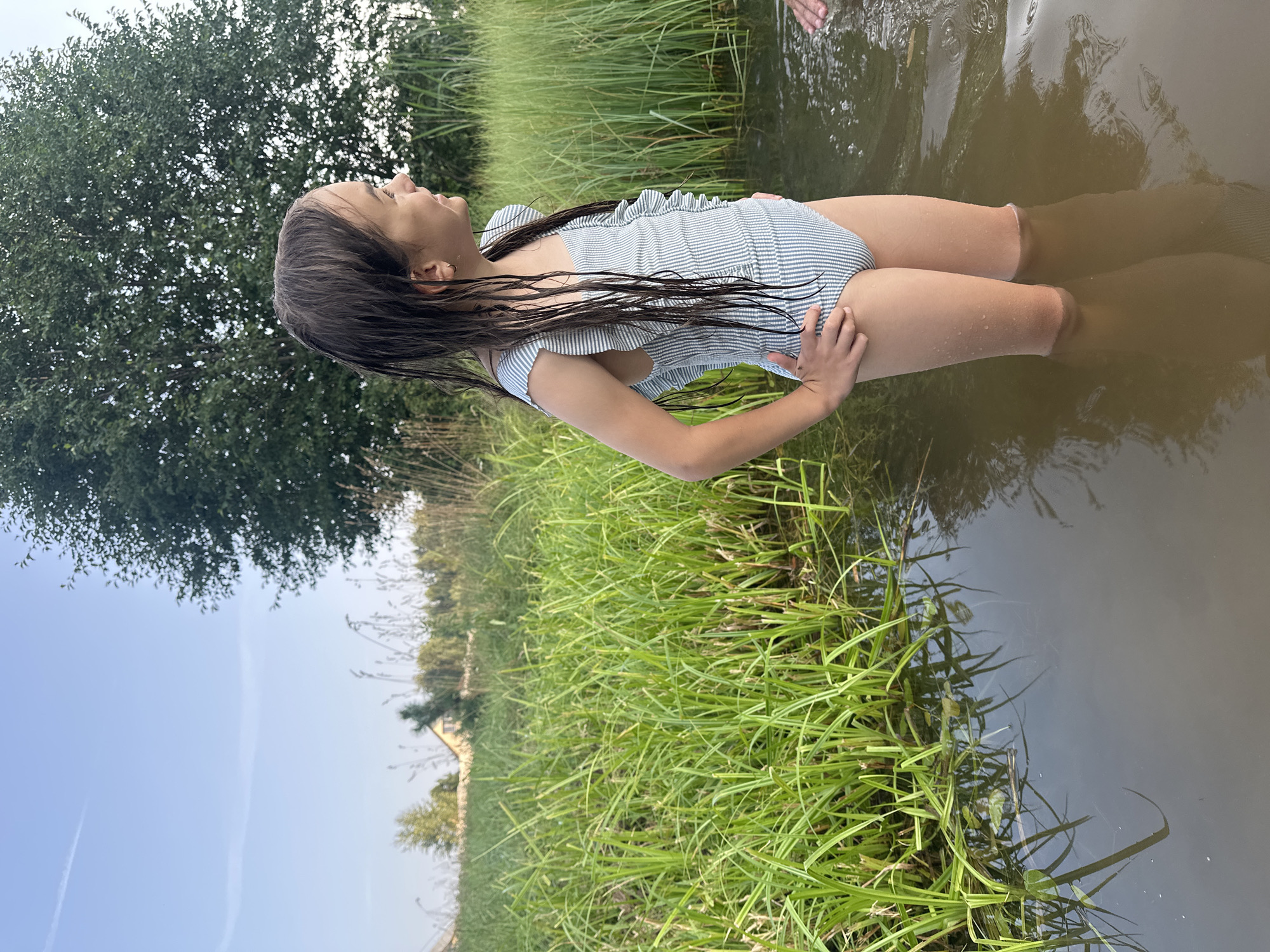 In this image, a young girl is standing in shallow water, with tall green grass surrounding her. She is wearing a striped swimsuit with ruffles on the shoulders and along the neckline. Her dark hair is wet and hangs down past her shoulders. She is looking upwards and to the left, with one hand on her hip and the other resting on her side. The water is murky and reflects the sky and surrounding vegetation. In the background, there is a large green tree on the left and more tall grass on the right. The sky is a pale blue with some faint wispy clouds. The overall impression is that of a warm, sunny day in a natural outdoor setting, likely a pond or lake. The girl appears relaxed and content. There is no visible text in the image. The location context suggests this is Kharnavy, Russia.