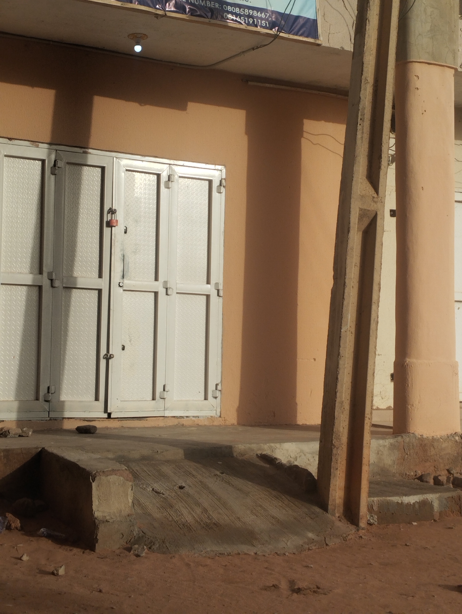 The image depicts a closed commercial building in Gombe, Nigeria. The building is peach-colored with white metal doors, secured with a padlock. A concrete ramp leads to the entrance. A concrete utility pole stands in the foreground. The ground is covered in reddish-brown soil and small debris.