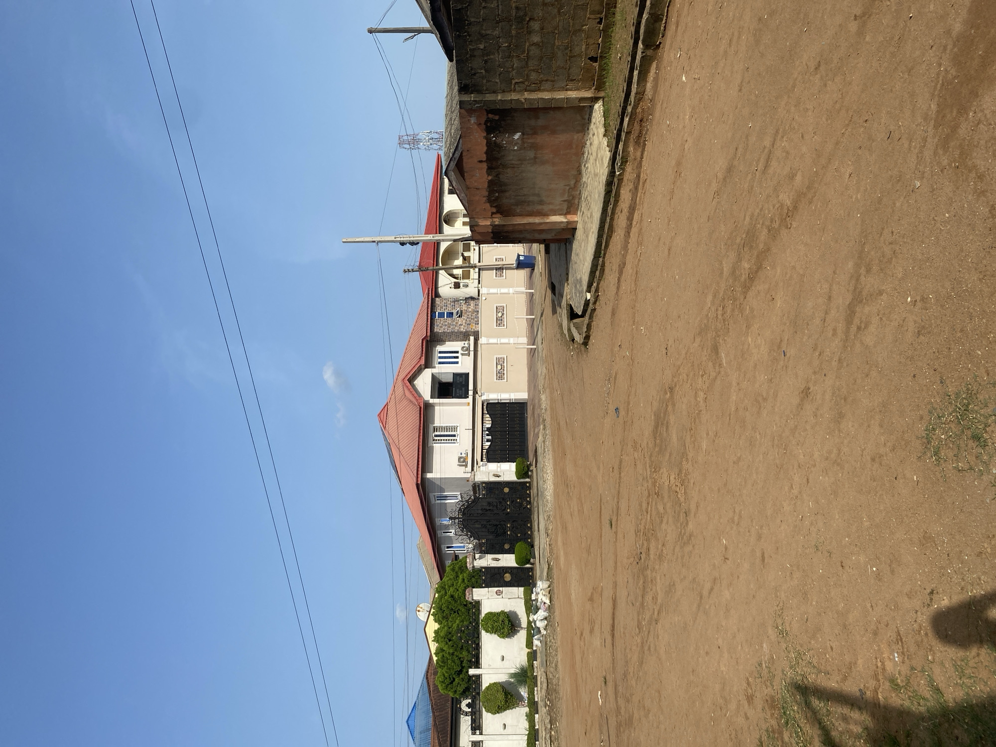 The image shows a street scene in Idimu, Nigeria. The road is unpaved, consisting of brown dirt. The right side of the image shows a partial view of a building constructed of brown bricks. In the background, there are two houses visible. One is cream-colored with a red roof, and a black gate. The other building is partially visible, with a blue roof. Power lines run across the top left of the image against a clear blue sky.