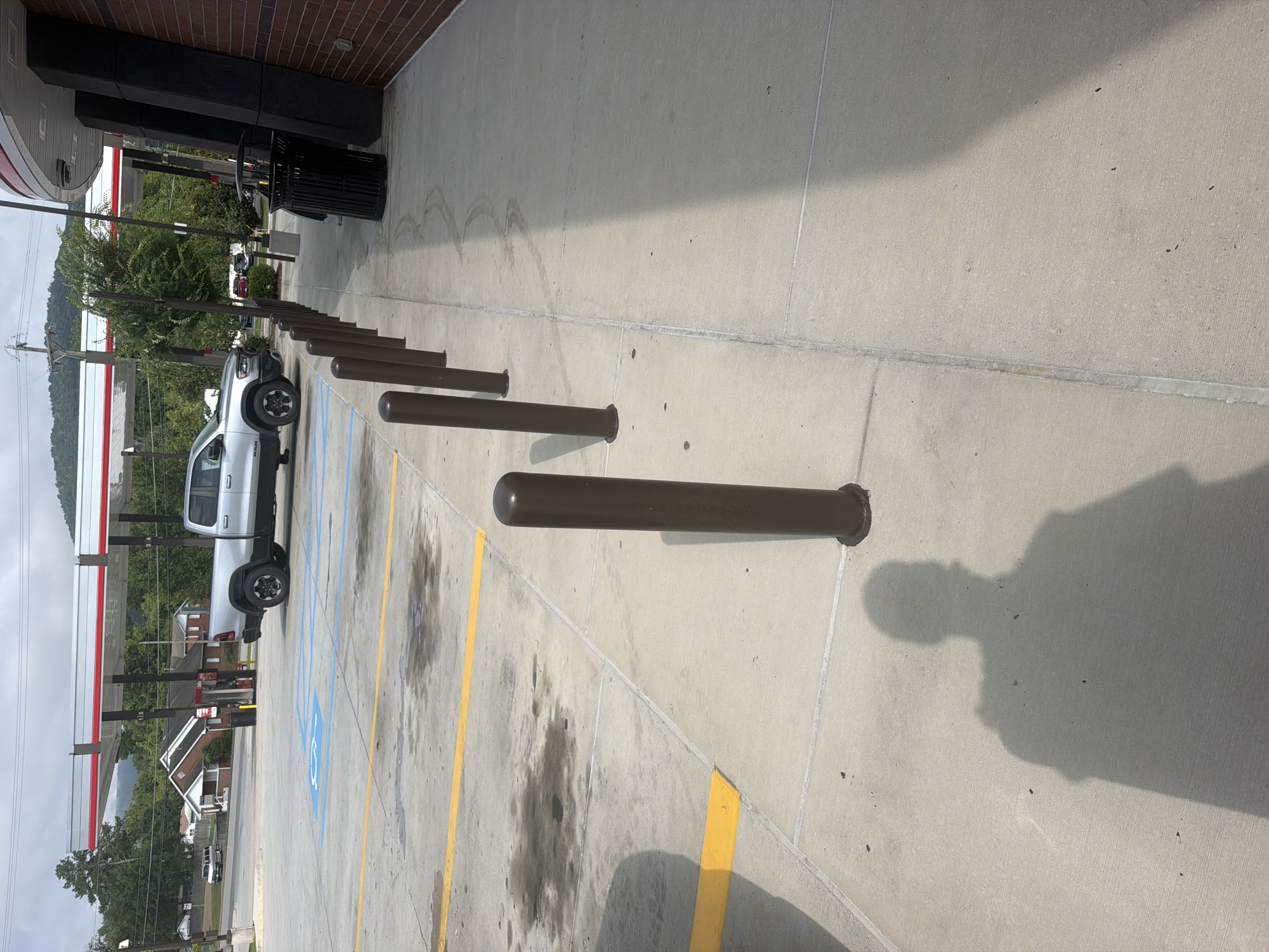 The image shows a sidewalk outside a gas station in Chattanooga, Tennessee. In the background, a silver pickup truck is parked at a gas pump under a red and white canopy. The sidewalk is lined with dark brown bollards. A black trash can is visible near the entrance of the building. The ground is concrete and shows some oil stains. The shadow of a person is cast on the sidewalk. The sky is cloudy, and trees and buildings are visible in the distance.