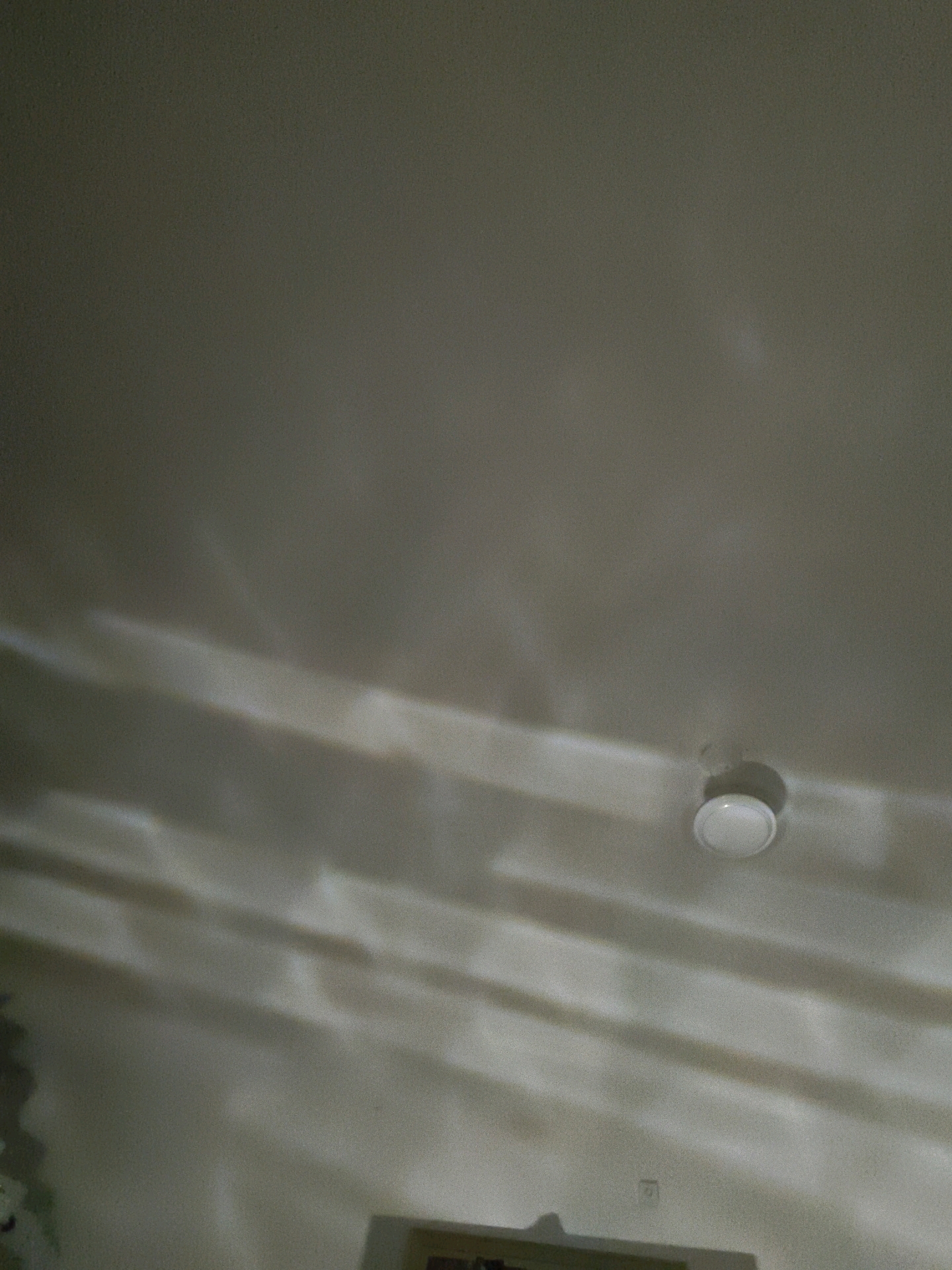 The image shows a close-up, angled shot of a textured ceiling in Mohali, India. The ceiling is a neutral, light color and appears to be painted drywall. Several horizontal bands of light and shadow are cast across the surface, suggesting illumination from a source outside the frame, possibly due to window blinds or other textured light-filtering elements. A white, circular light fixture or smoke detector is mounted on the ceiling in the lower right quadrant of the image. There is a small, square electrical outlet visible on the wall in the lower middle of the image. The scene is indoors and appears to be a domestic or office environment.