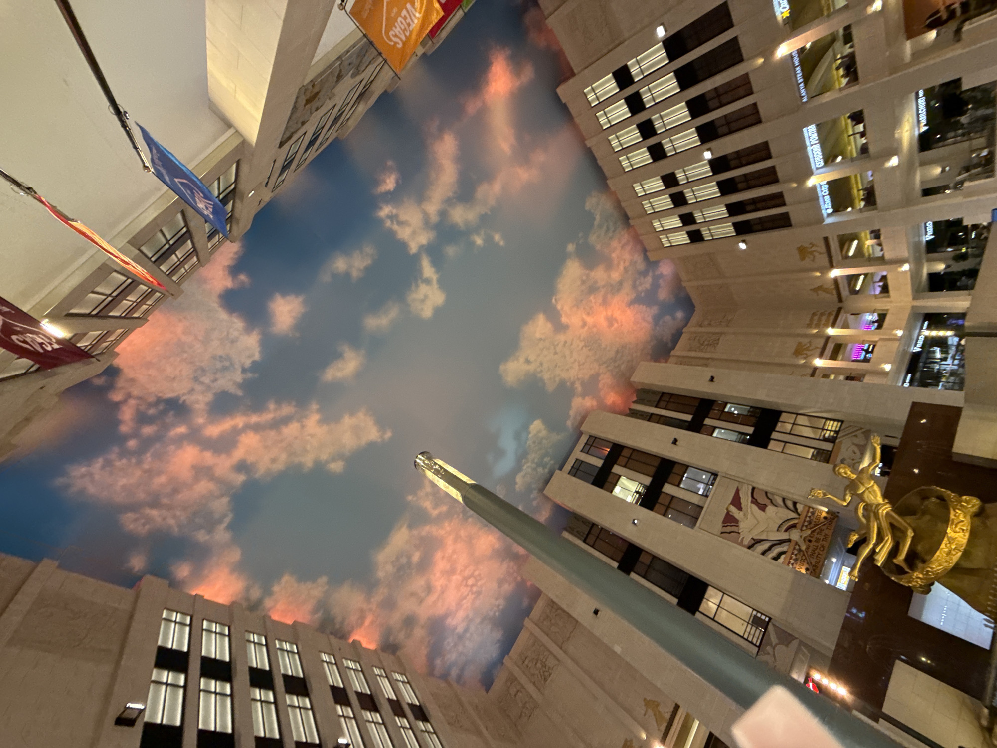 The image shows an interior view of a shopping mall, likely in Khimki, Russia, with a high ceiling painted to resemble a sunset sky with clouds. The architecture features multi-story buildings with large windows and decorative elements on the facade, appearing to be in an Art Deco style. Several flags are hanging from the ceiling. On a lower level, a golden statue of a person or mythological figure is visible, along with various shops and restaurants with illuminated signage. The lighting suggests it is either evening or the mall's artificial lighting is creating a dramatic effect.