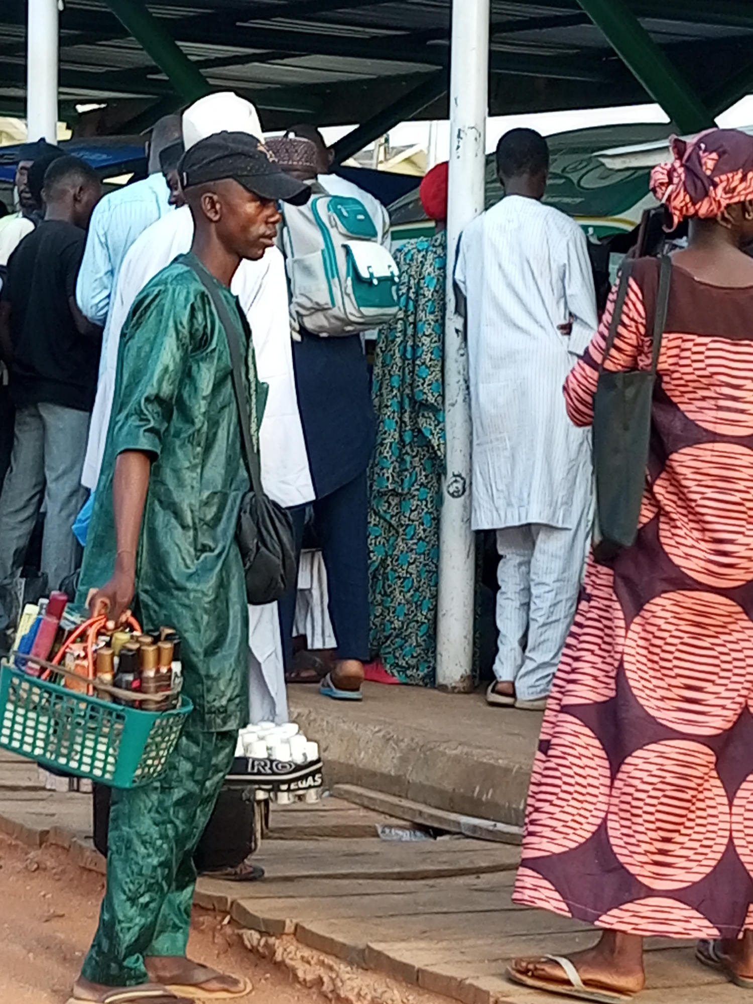 The image depicts a street scene in Gombe, Nigeria, likely a market or a busy thoroughfare. In the foreground, a man wearing a green traditional garment and a black baseball cap carries a green basket filled with small bottles and containers, possibly perfumes or medications. He is also wearing a dark shoulder bag. To his right, a woman in a colorful patterned dress and headscarf is visible, also carrying a dark bag. Other individuals in the background are dressed in various traditional and modern attire, suggesting a diverse crowd. The environment appears to be outdoors, with a covered structure overhead, indicated by the presence of support beams and a roof. The ground is paved with wooden planks and some dirt. The overall atmosphere seems to be that of a bustling commercial area.