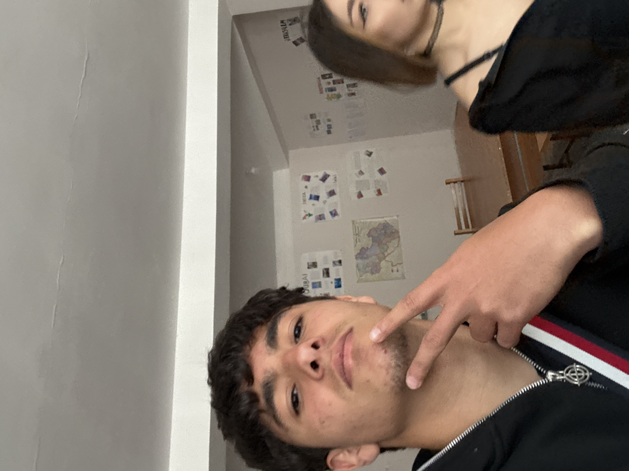 A medium, slightly distorted selfie depicts two individuals in an interior space. The main subject is a young, brown-haired male with stubble pointing at his cheek. He is wearing a black jacket with red and white stripes and silver zipper. A female with brown hair and a black top is partially visible in the background. The background includes a wall with several posters, some possibly showcasing images of various cities including Dubai and Jerusalem. A map and white radiator are also visible. While the location cannot be determined based on this image, there is nothing to suggest it is located in Yerevan, Armenia.