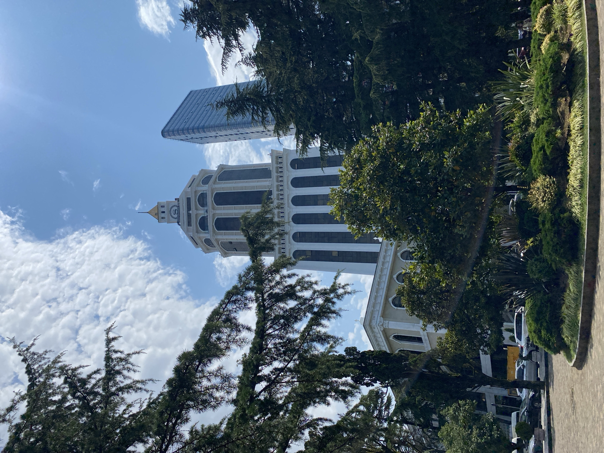 The image shows a low-angle view of the Batumi Boulevard area in Batumi, Georgia. In the background, two prominent buildings are visible: a tall, white, art-deco style tower with a golden spire and clock face, and a modern, glass-fronted skyscraper. Lush green trees and manicured bushes frame the foreground, partially obscuring the buildings and suggesting a park-like setting. The sky above is blue with scattered white clouds, and sunlight casts a lens flare across the scene. In the middle ground, cars are parked and driving on a street, indicating an urban environment. The overall impression is one of a sunny day in a coastal city, blending historical and contemporary architecture with natural elements.