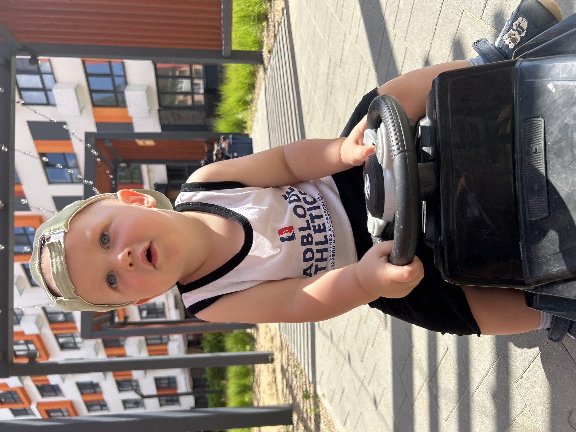 A young Caucasian child, wearing a light green baseball cap, a white tank top with "ADBLO ATHLETIC" printed on it, and black shorts, sits on a black toy vehicle. The child's hands are on the steering wheel, and they are looking upwards and to the left with a surprised expression. The child is wearing dark blue shoes with a teddy bear decal. The background features a modern residential building with orange and white accents, along with green foliage and a paved walkway. The scene appears to be an outdoor area in a city.