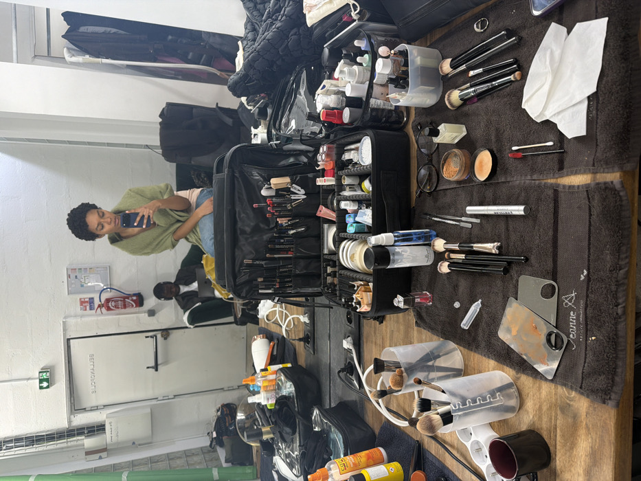 The image depicts a professional makeup station, likely in a studio or backstage dressing room, located in the 3rd arrondissement of Paris, France (confirmed by "75003 PARIS FRANCE" text on a towel).
In the foreground, a wooden table is covered with dark brown towels, meticulously displaying a comprehensive array of makeup tools and products. A large, open, multi-compartment black makeup case occupies the center, filled with numerous brushes, palettes, and cosmetic containers. Around it are smaller clear containers holding additional brushes, various makeup bottles (including setting sprays and foundations), powders, mascaras, lip products, and application tools. A black coffee mug, a white power strip, a pair of black-rimmed glasses, and used makeup palettes are also visible on the table.
The background of the image is a reflection in a large mirror. A young woman with short, curly dark hair, wearing a light green shawl over a pink top and blue jeans, is seated, holding a smartphone and seemingly taking a selfie or examining her reflection. Behind her, also in the reflection, a young man with dark skin, wearing headphones, is seated on a dark green couch, focused on a laptop. The reflected wall is white, featuring a green emergency exit sign, a red fire extinguisher, and a white door with reversed text. To the far right, clothing racks draped with dark garments are visible. The scene portrays a moment of preparation and waiting in a professional environment.